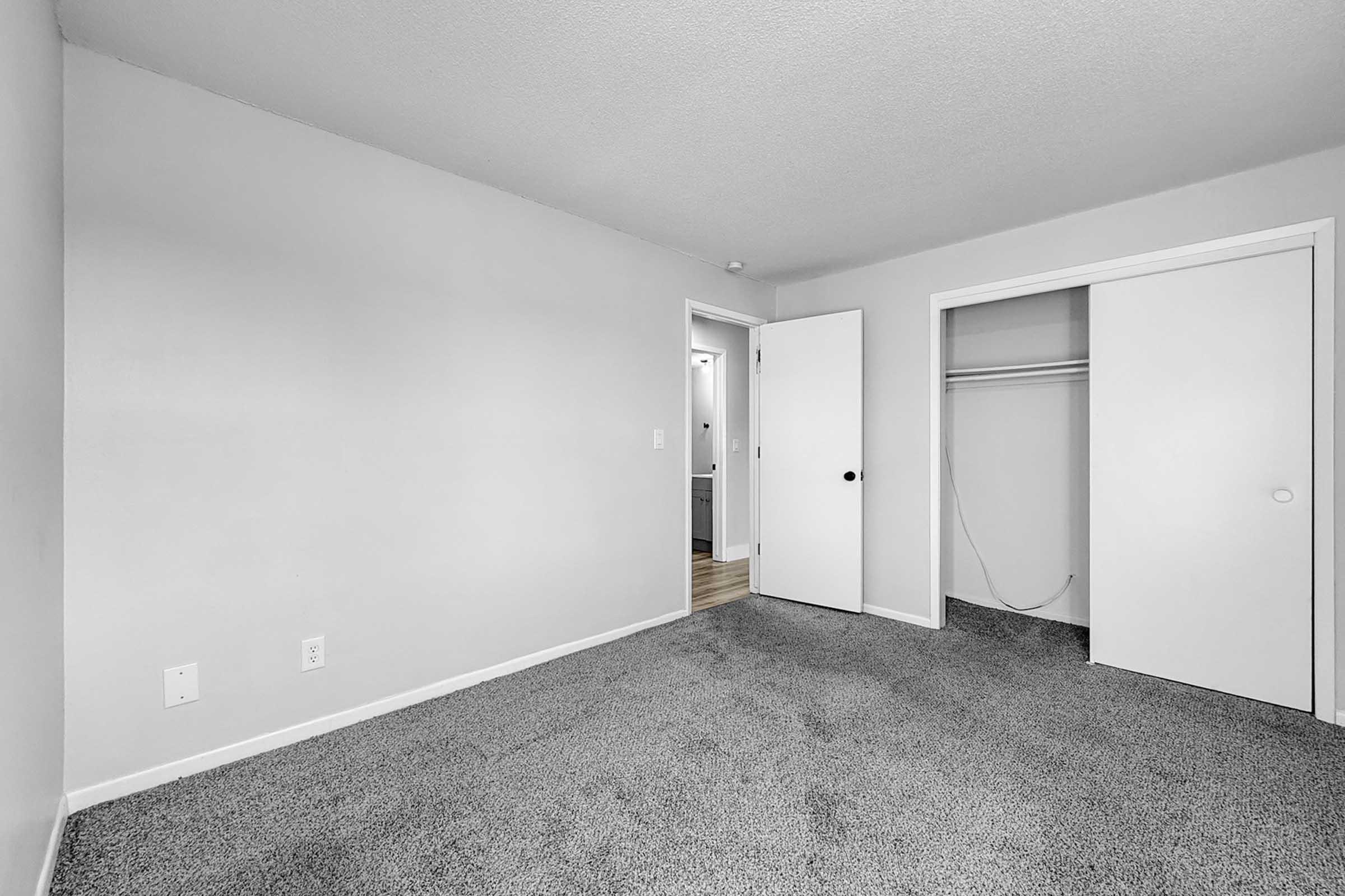 A sparsely furnished room with light gray walls and carpet. There is a closed closet on the right with double doors, and an open doorway leading to another room on the left. The space is well-lit and appears clean and spacious.