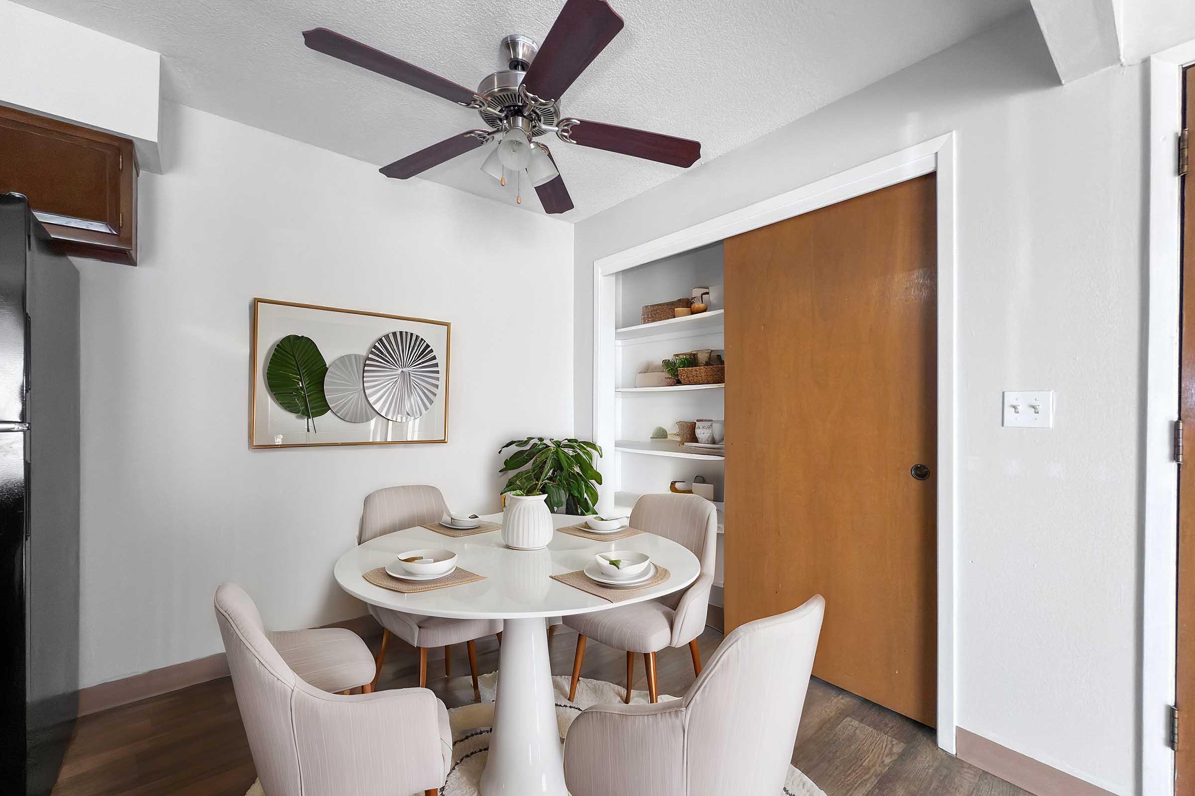 A cozy dining area featuring a small round table with four cream-colored chairs. The table is set with dishes and cutlery. There's a plant on the table and a wall-mounted shelf displaying decorative items. A ceiling fan and a wooden door complete the space, with light-colored walls for a bright atmosphere.