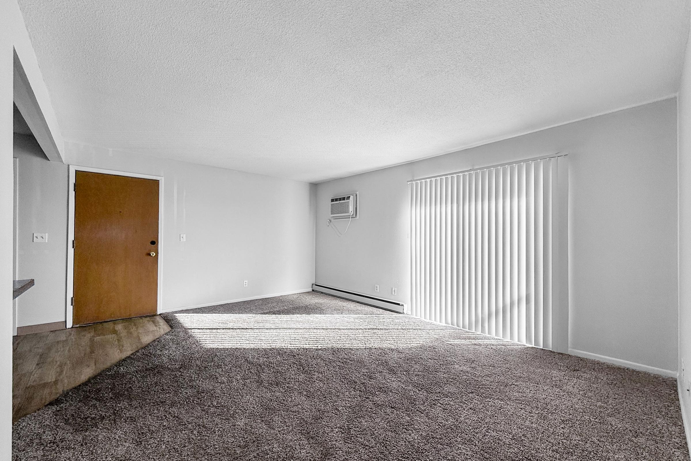 Spacious, empty living room with beige carpet, a wall-mounted air conditioning unit, and large windows covered with vertical blinds. Natural light streams in, highlighting the room's clean, neutral walls and simple decor. A wooden front door is visible on the left.