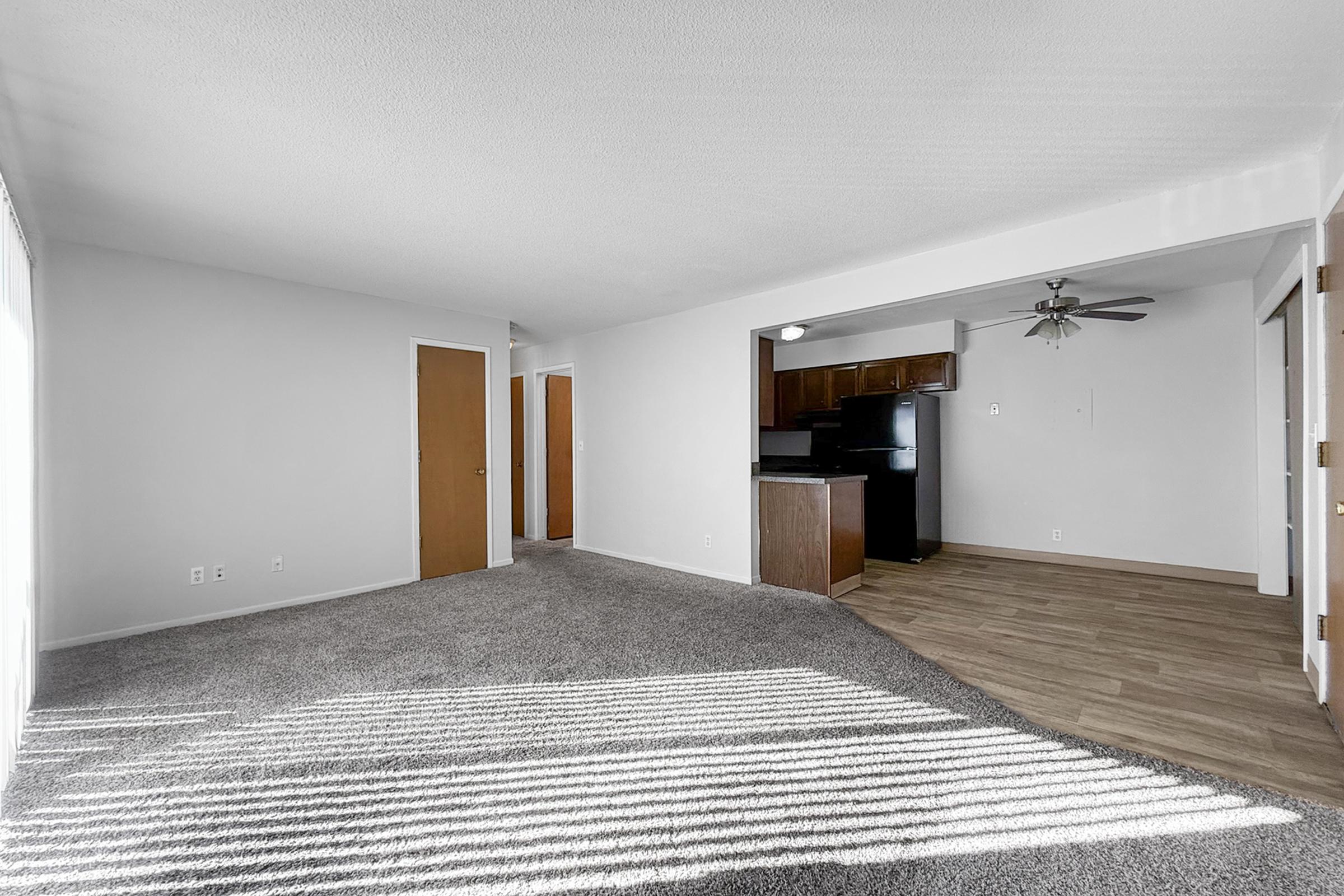 View of a spacious living area with light-colored carpet, a ceiling fan, and four doors leading to different rooms. The kitchen is partially visible on the right, featuring dark cabinets and appliances. Natural light streams in through a window, casting striped shadows on the floor.