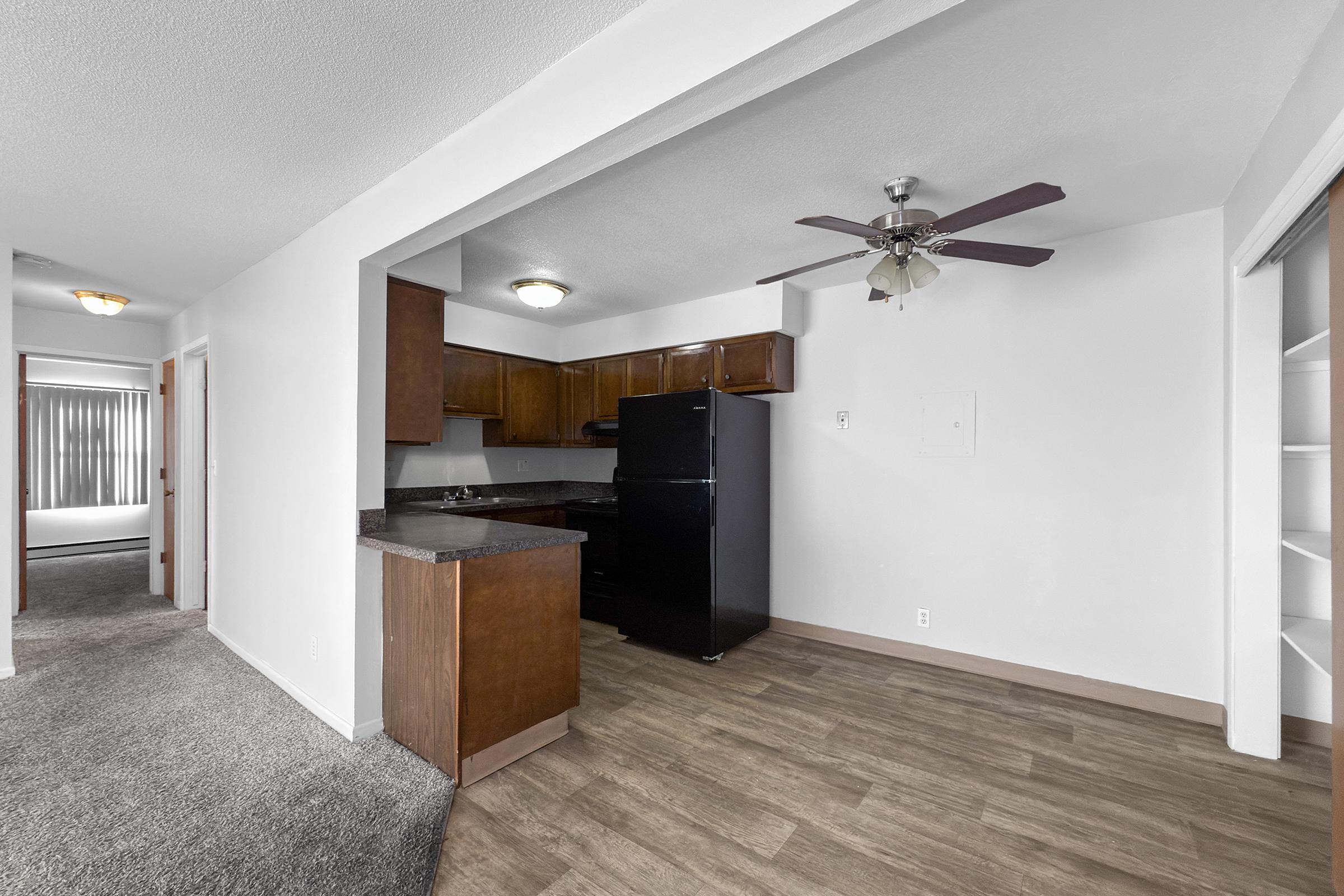 A view of a spacious kitchen area featuring dark wood cabinets, a black refrigerator, and a ceiling fan. The kitchen has a countertop extending into a living space, with a doorway leading to a separate room. The floor is covered with light-colored laminate, and there's carpet in the adjacent area.