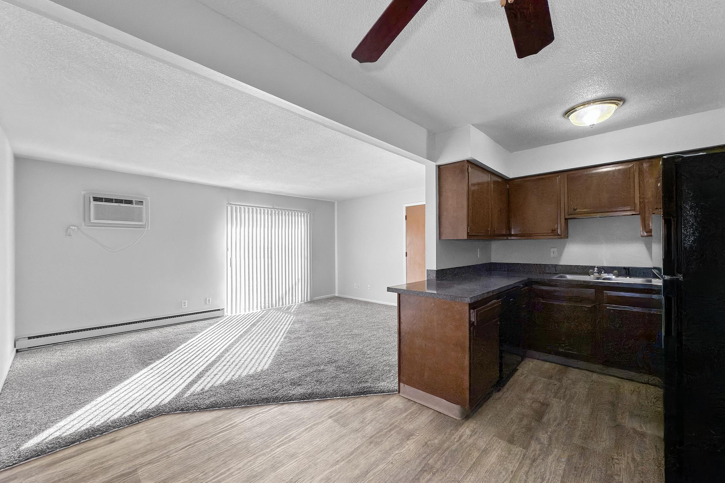 Open living area featuring a kitchen with dark wood cabinets and a black refrigerator. There is a window allowing natural light to enter, along with an air conditioning unit on the wall. The floor is a combination of carpet and laminate. Textured ceiling and ceiling fan visible.