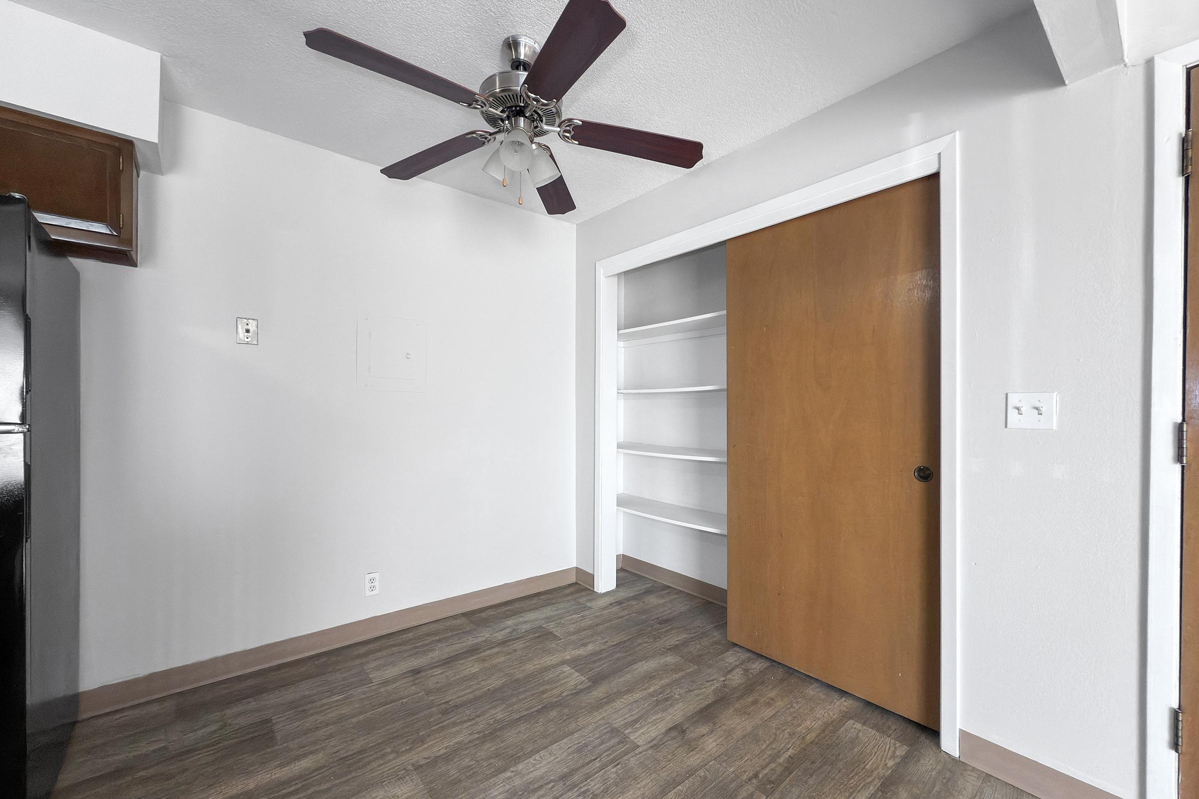 A small, simple room features a ceiling fan with dark wood blades, light-colored walls, and hardwood floors. There's a black refrigerator visible on the left, and to the right, a sliding wooden door leading to an open closet with shelves. The space is well-lit and unobstructed.
