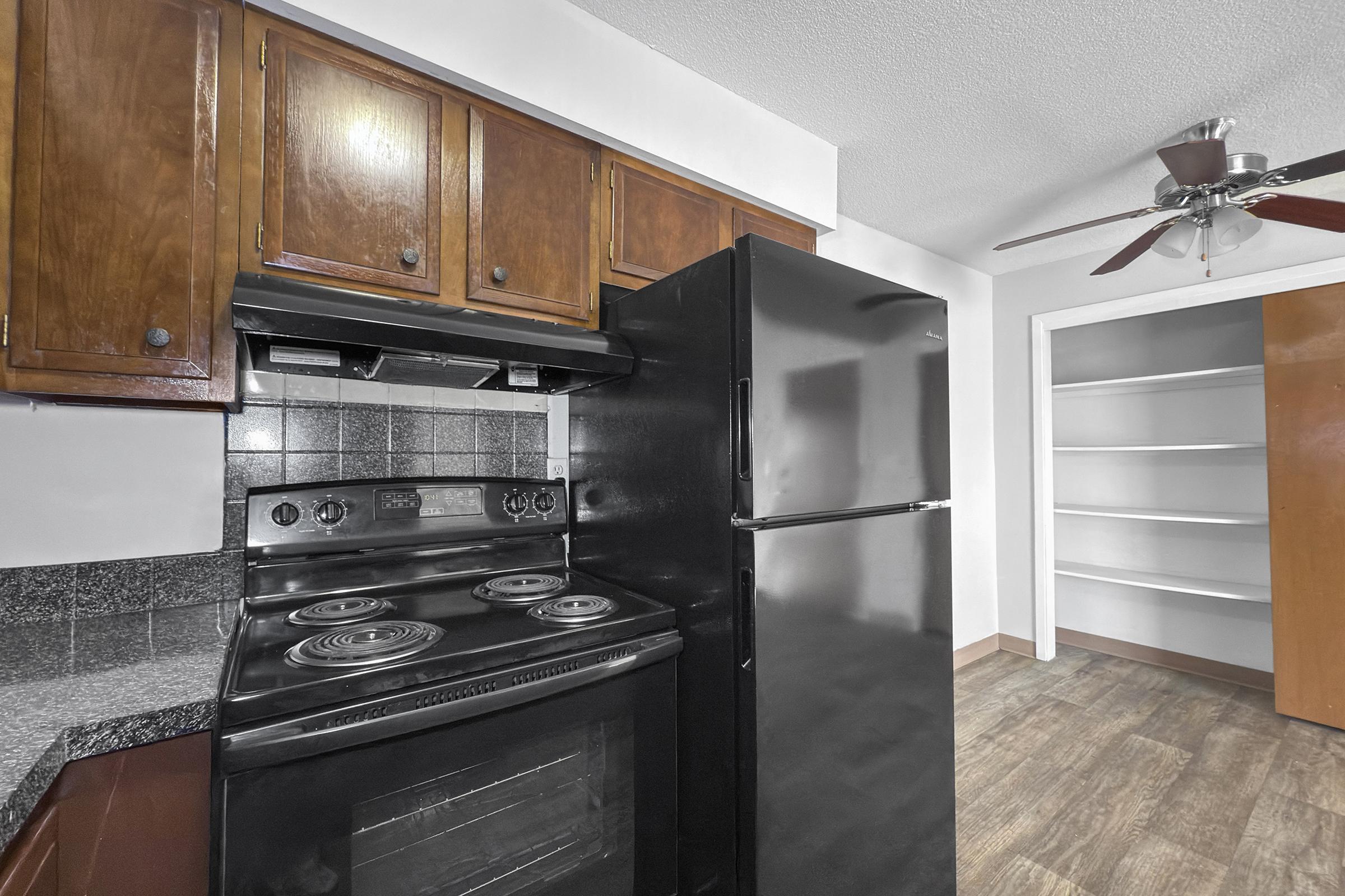 A modern kitchen featuring dark wood cabinetry, a black stove and refrigerator, and textured gray countertops. A ceiling fan is visible, and there is a spacious pantry or closet with shelves in the background. The flooring is a light wood look, enhancing the contemporary feel of the space.