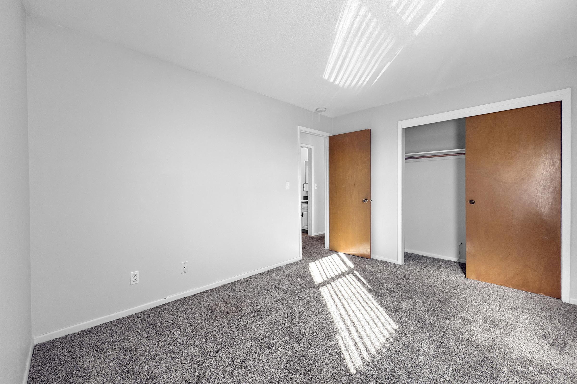 A bright, minimally furnished room with gray carpet and light-colored walls. Sunlight streams through a window, casting stripes on the floor. The room features two wooden doors, one leading to a closet and the other possibly to another space. The overall appearance is clean and simple.