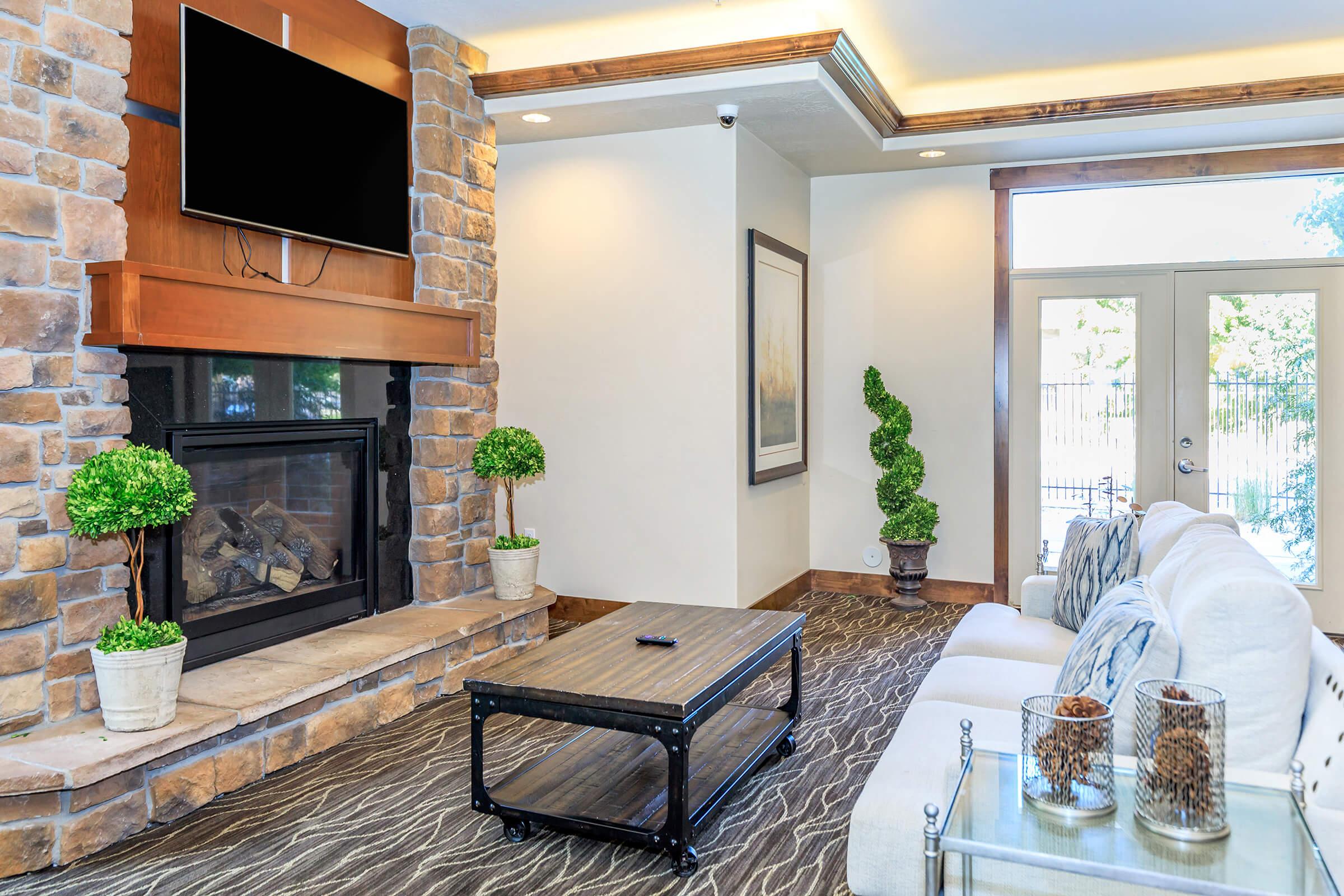 A cozy living room featuring a stone fireplace with a mounted television above it. A stylish coffee table is in front of a comfortable white couch. Potted plants add a touch of greenery, and large windows offer a view of an outdoor area. The decor is modern and inviting, with warm lighting throughout the space.