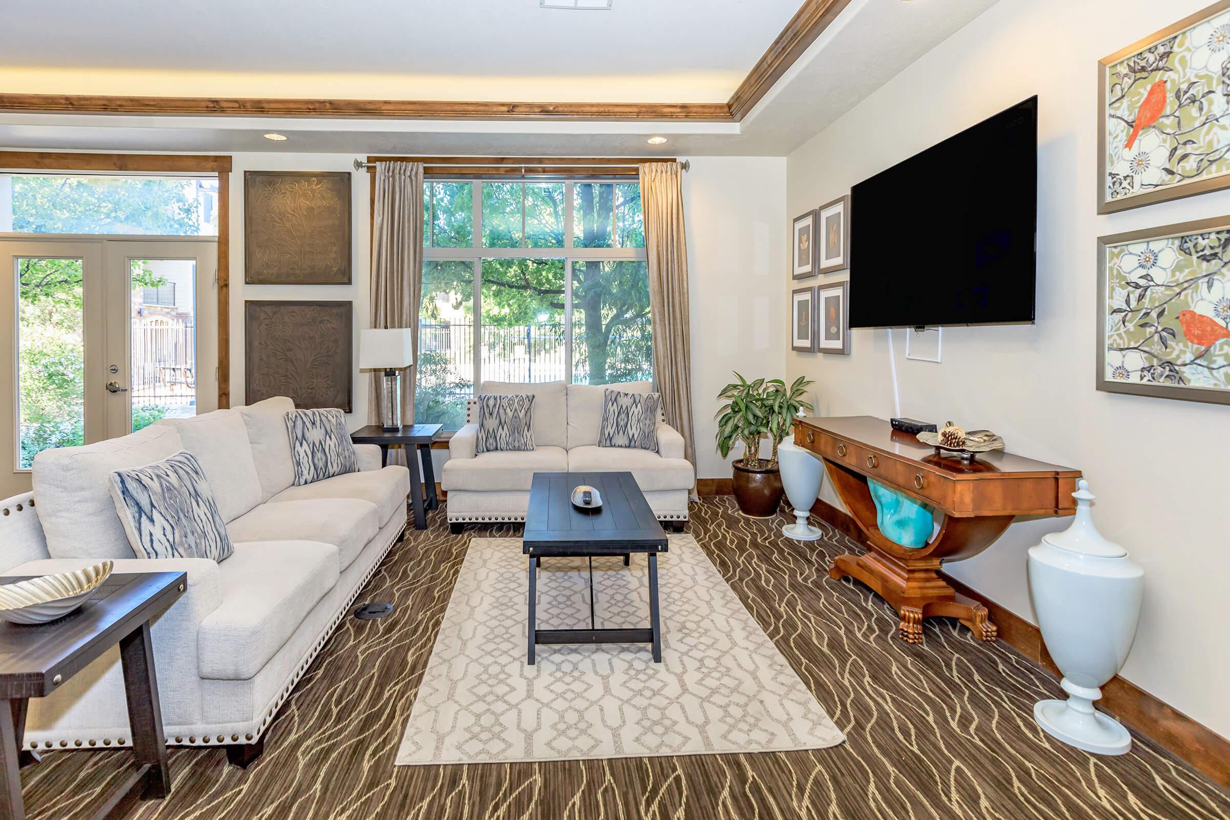 A cozy living room featuring a light-colored sofa and armchair, a central coffee table, a large television mounted on the wall, decorative artwork, and potted plants. Large windows allow natural light, and the space has a warm, inviting atmosphere with a patterned rug.