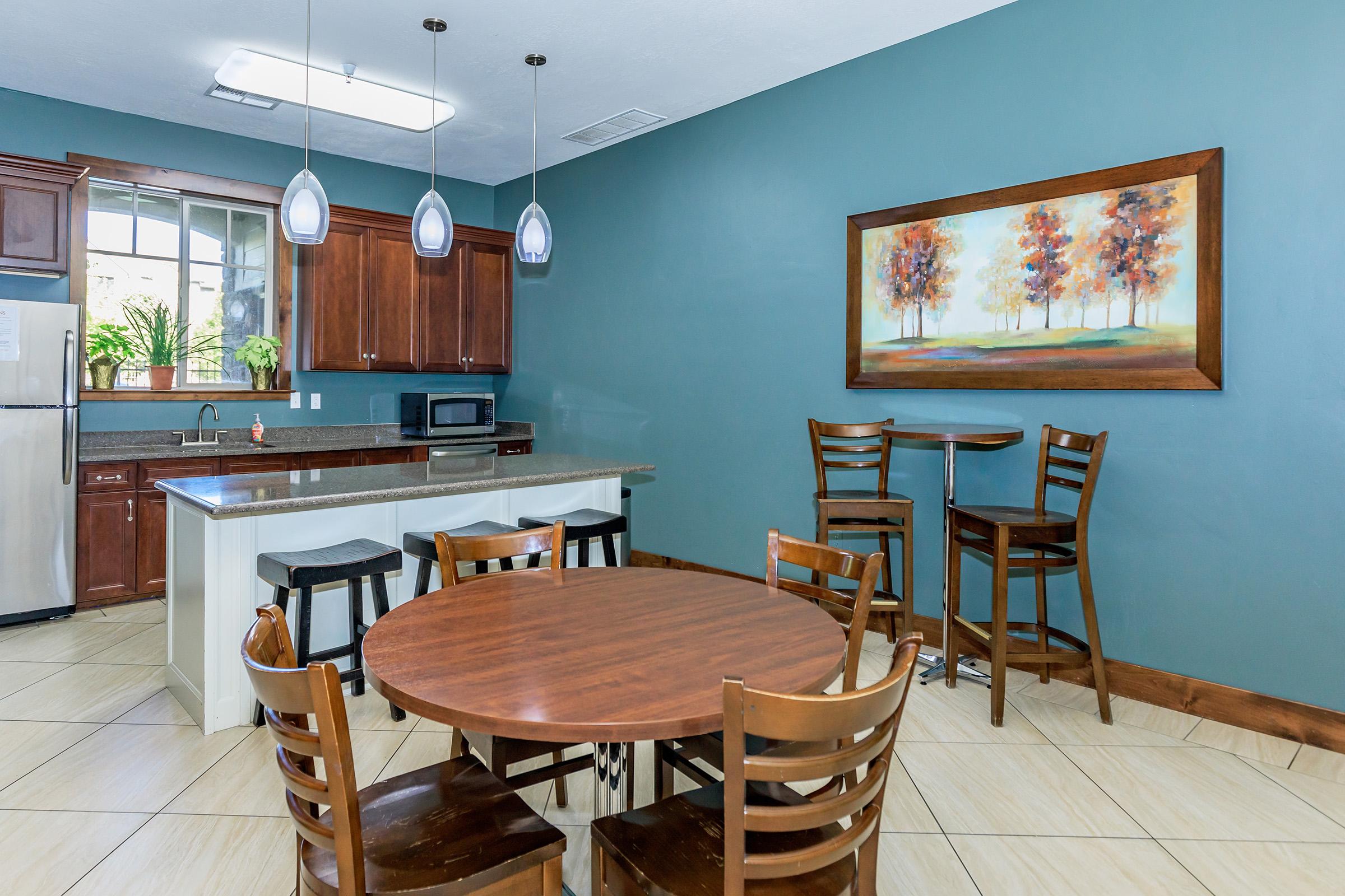 A modern kitchen with dark wood cabinets and stainless steel appliances. There is a circular wooden dining table with six chairs, and two high stools at a small counter. The walls are painted teal, and a landscape painting with trees hangs on the wall. Natural light comes in from a window.