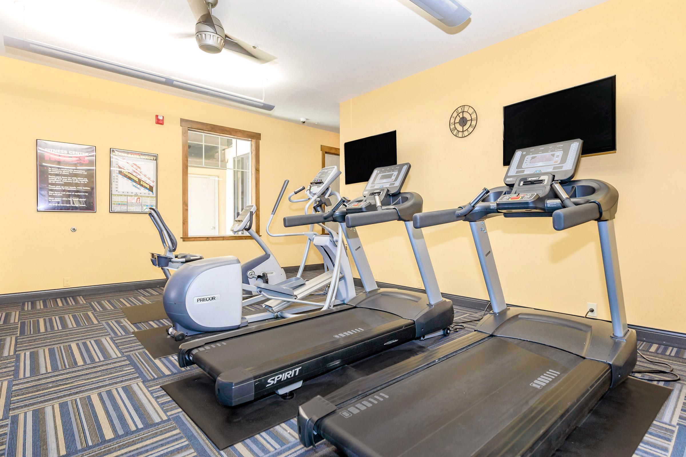 A fitness room featuring several exercise machines including treadmills and an elliptical. The walls are painted yellow, and there are two wall-mounted televisions. The floor is carpeted in a blue geometric pattern, and motivational posters are displayed on the walls.