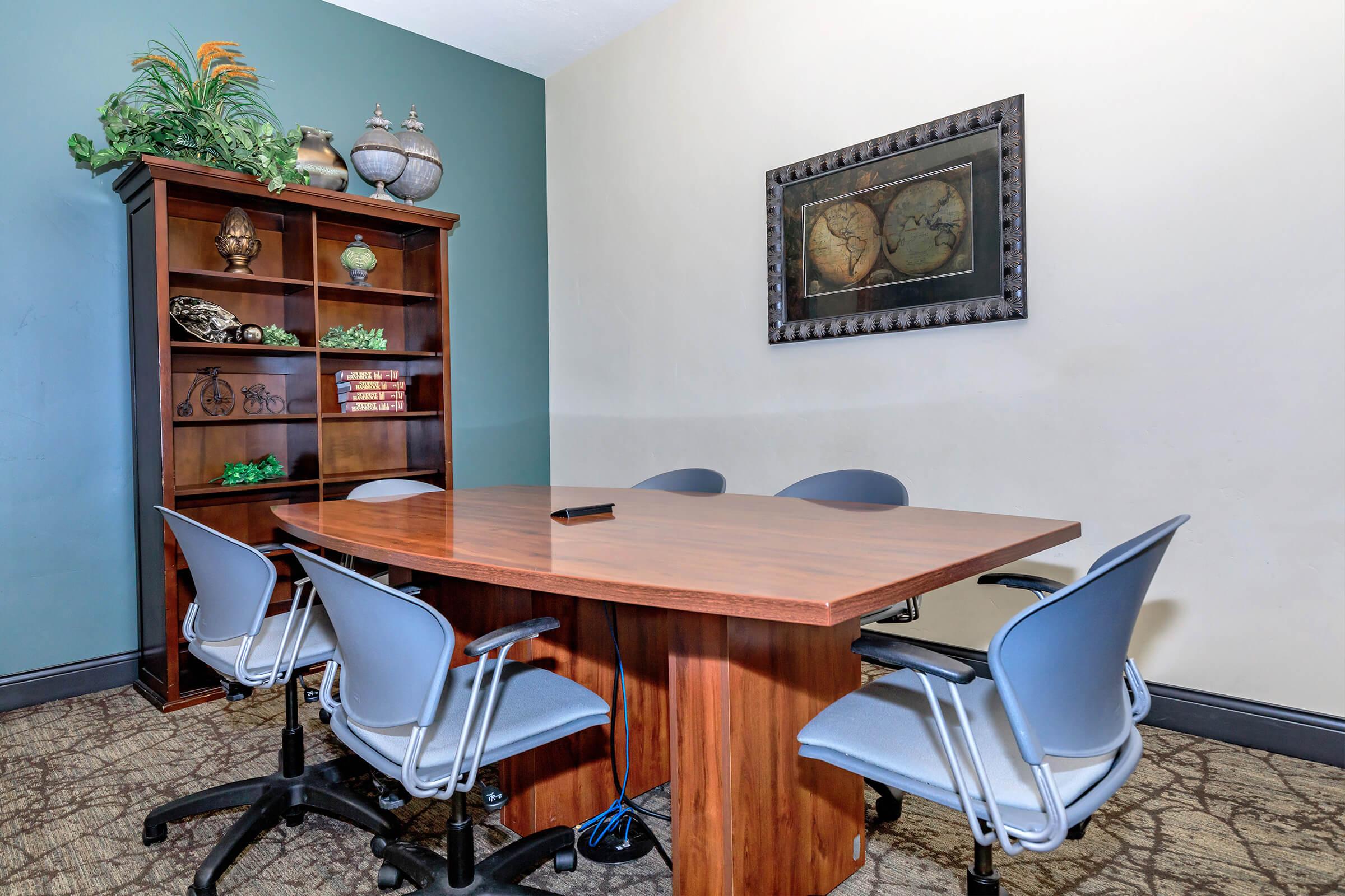 A clean and modern meeting room featuring a wooden table surrounded by four gray chairs. There's a bookshelf against the wall displaying decorative items and books, and a framed artwork above the table. The walls are painted in soft colors, creating a professional atmosphere.