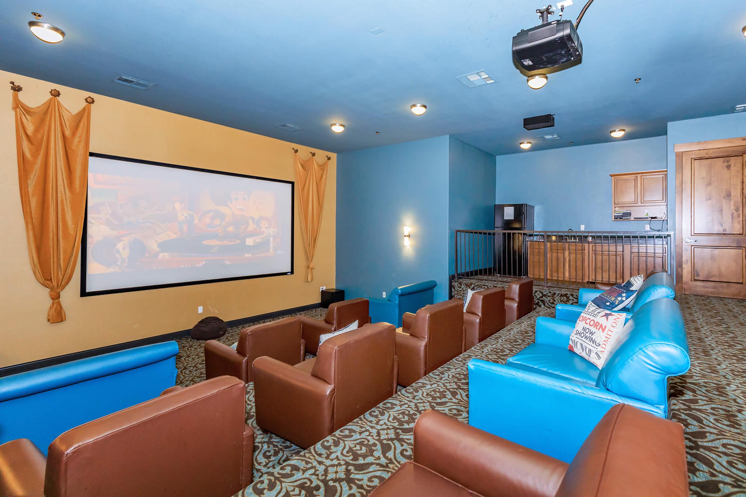 Home theater room featuring a large screen, brown and blue seating, decorative curtains, and a kitchenette area in the background. The walls are painted in a light blue shade, creating a cozy atmosphere for viewing movies or presentations.