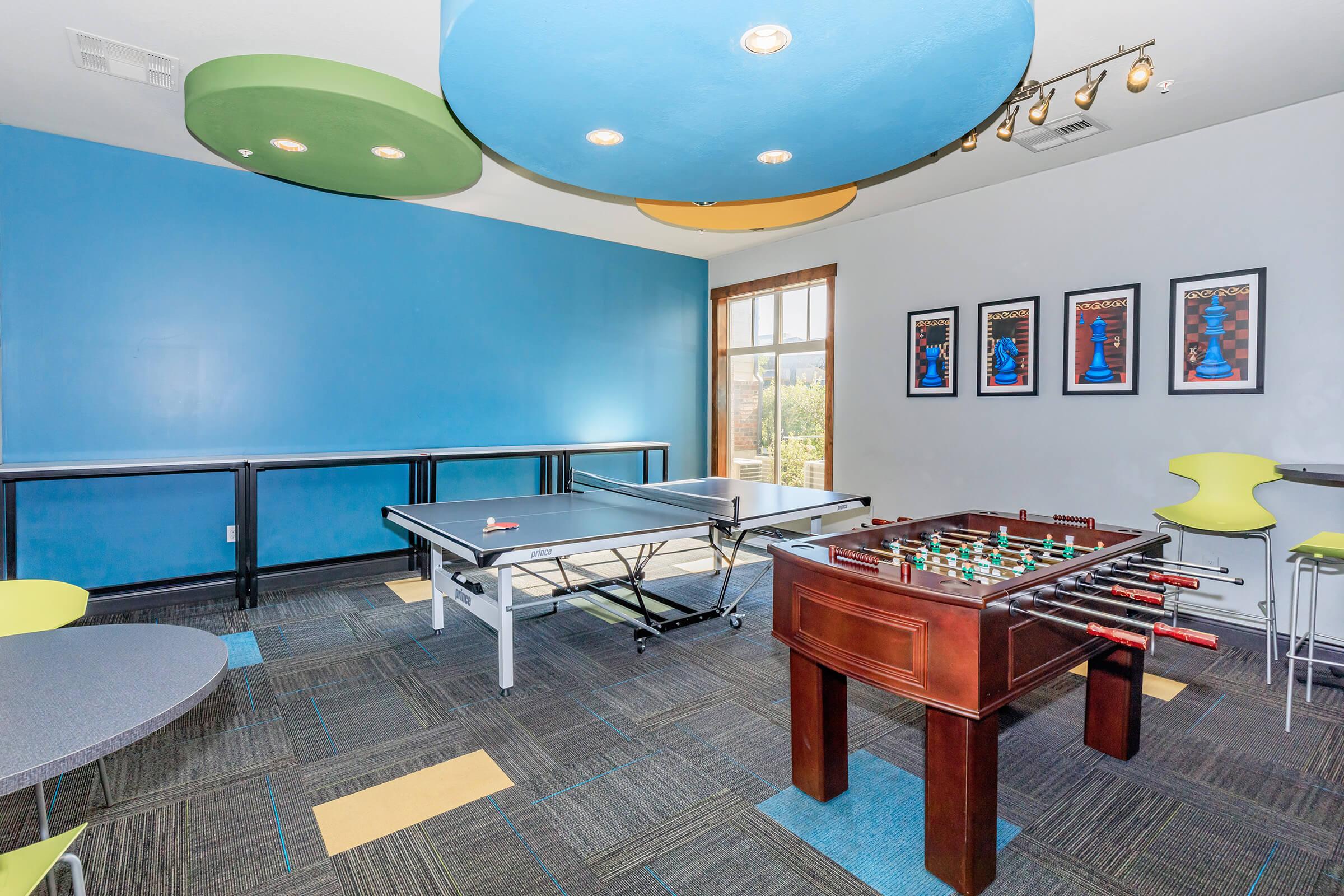 A recreational room featuring a ping pong table and a foosball table. The walls are painted blue, with colorful circular ceiling fixtures in green and orange. There are modern seating areas arranged around the room, and framed artwork is displayed on the wall. Bright, vibrant decor creates an inviting atmosphere.