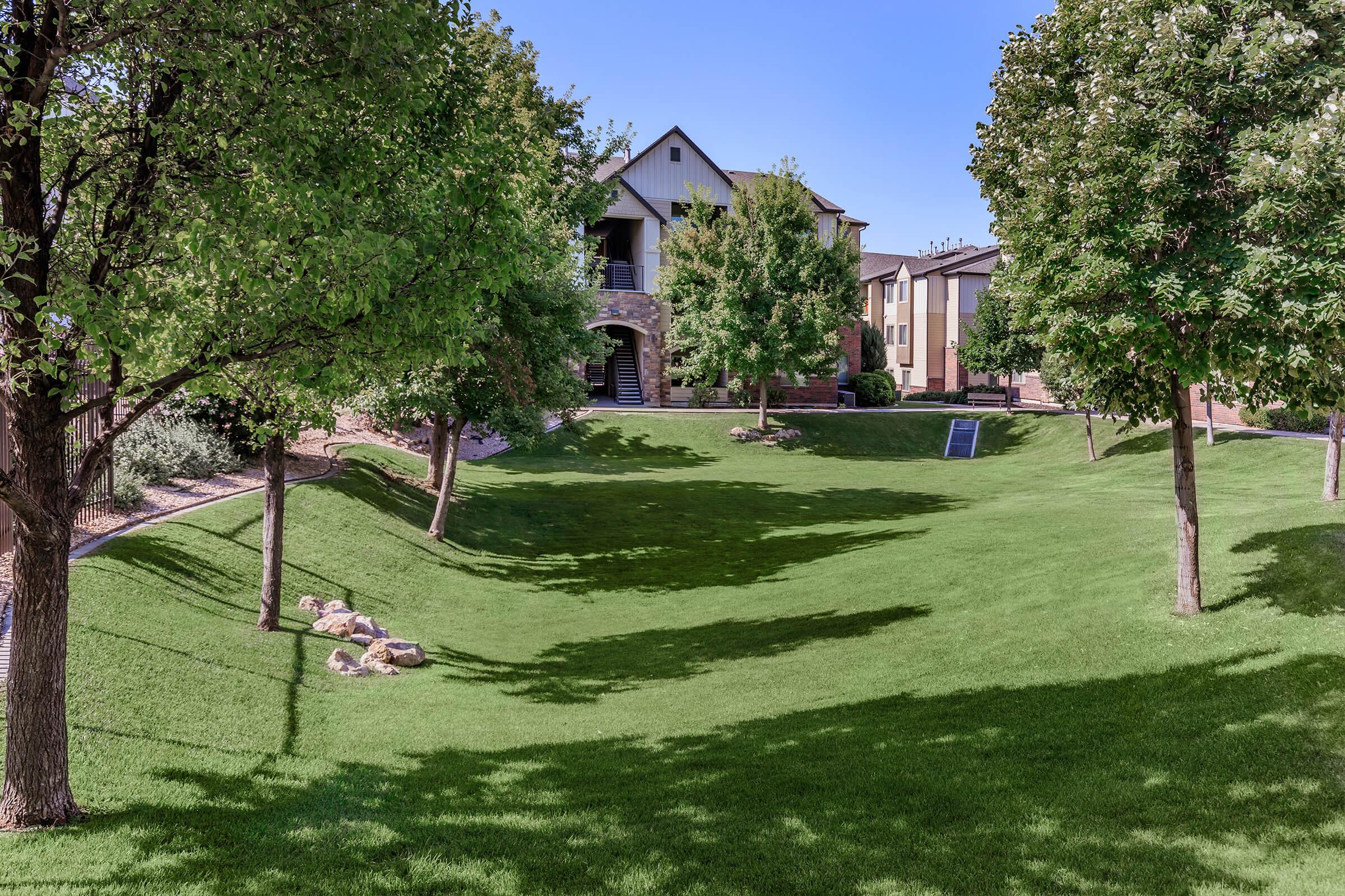 Lush green landscape featuring a gently sloping lawn with well-maintained grass and trees, set against a backdrop of residential buildings. The scene is bright and sunny, showcasing the natural beauty of the outdoor space. Shadows from the trees create a pleasant interplay of light and shade on the ground.