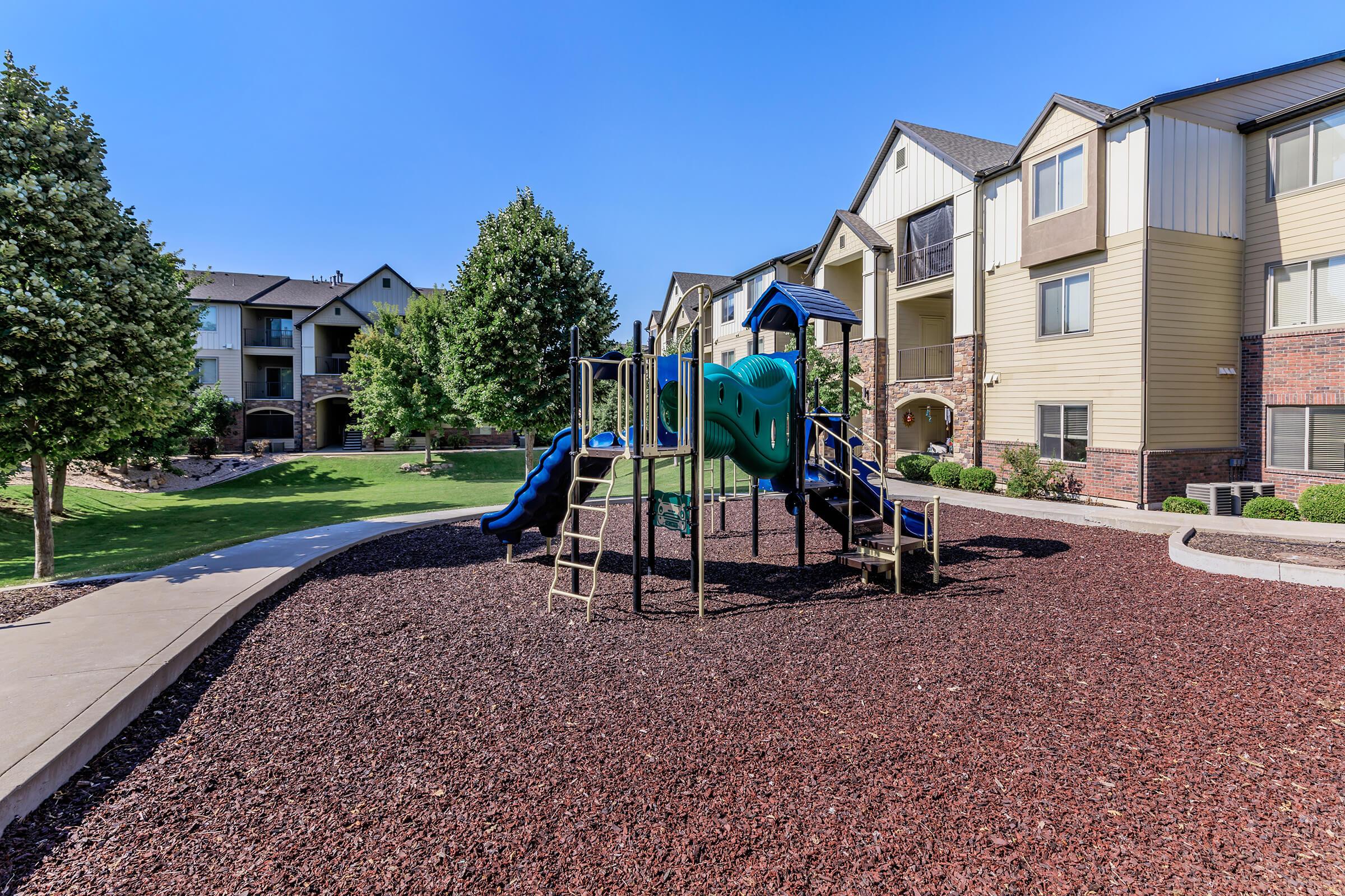 A colorful children's playground featuring a slide and climbing structure, surrounded by landscaped greenery and trees. Apartment buildings are visible in the background under a clear blue sky. The ground is covered with red mulch, creating a playful environment.