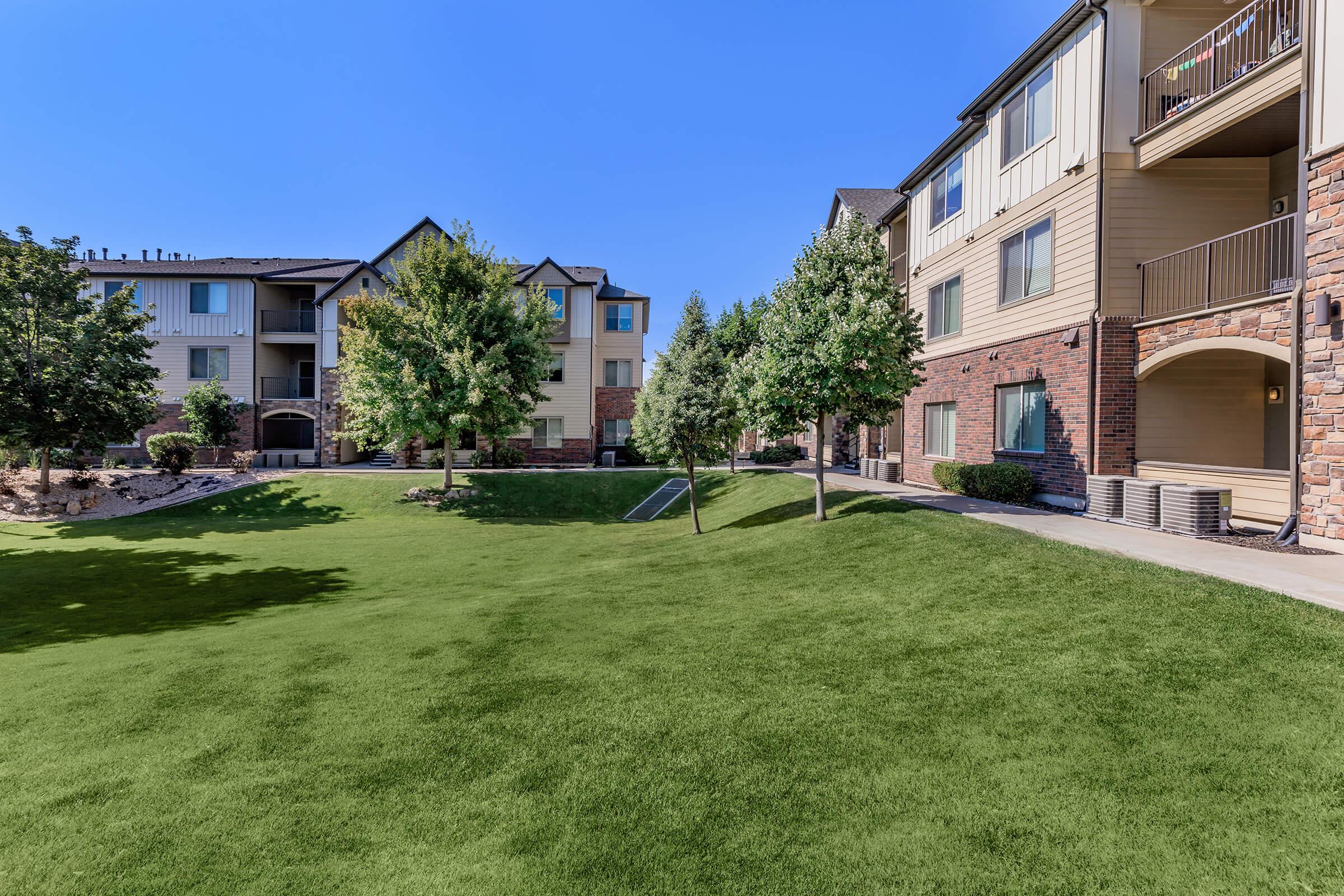 A well-maintained residential complex featuring multiple two- and three-story buildings surrounded by lush green lawns and trees. The clear blue sky enhances the sunny and inviting atmosphere of the outdoor space, which includes pathways and landscaped areas for residents to enjoy.