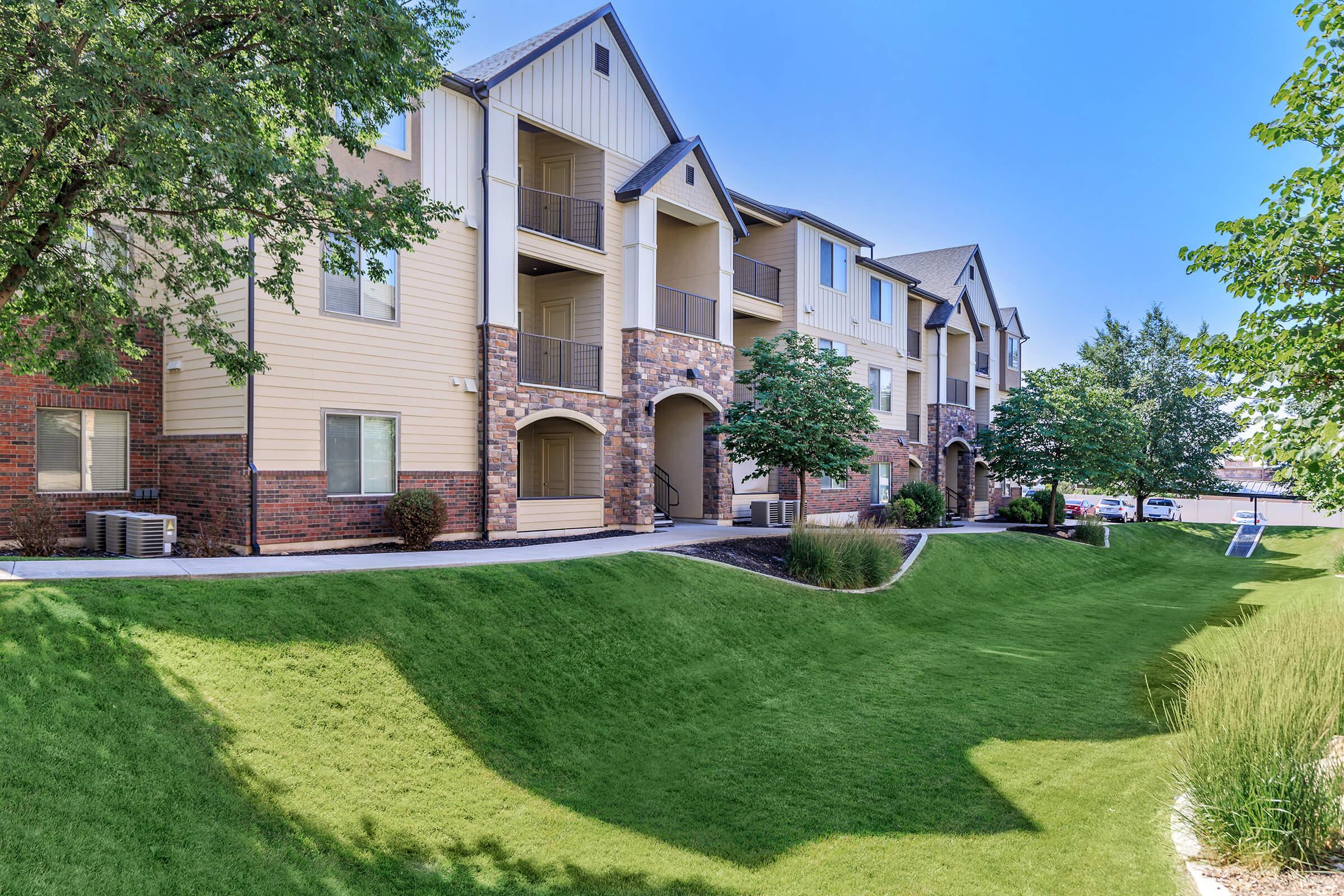 A modern apartment community featuring multiple buildings with a mix of brick and siding exteriors. The landscape includes well-maintained green lawns, trees, and shrubs, creating a welcoming outdoor environment. Balconies and patios enhance the residential appeal of the buildings.