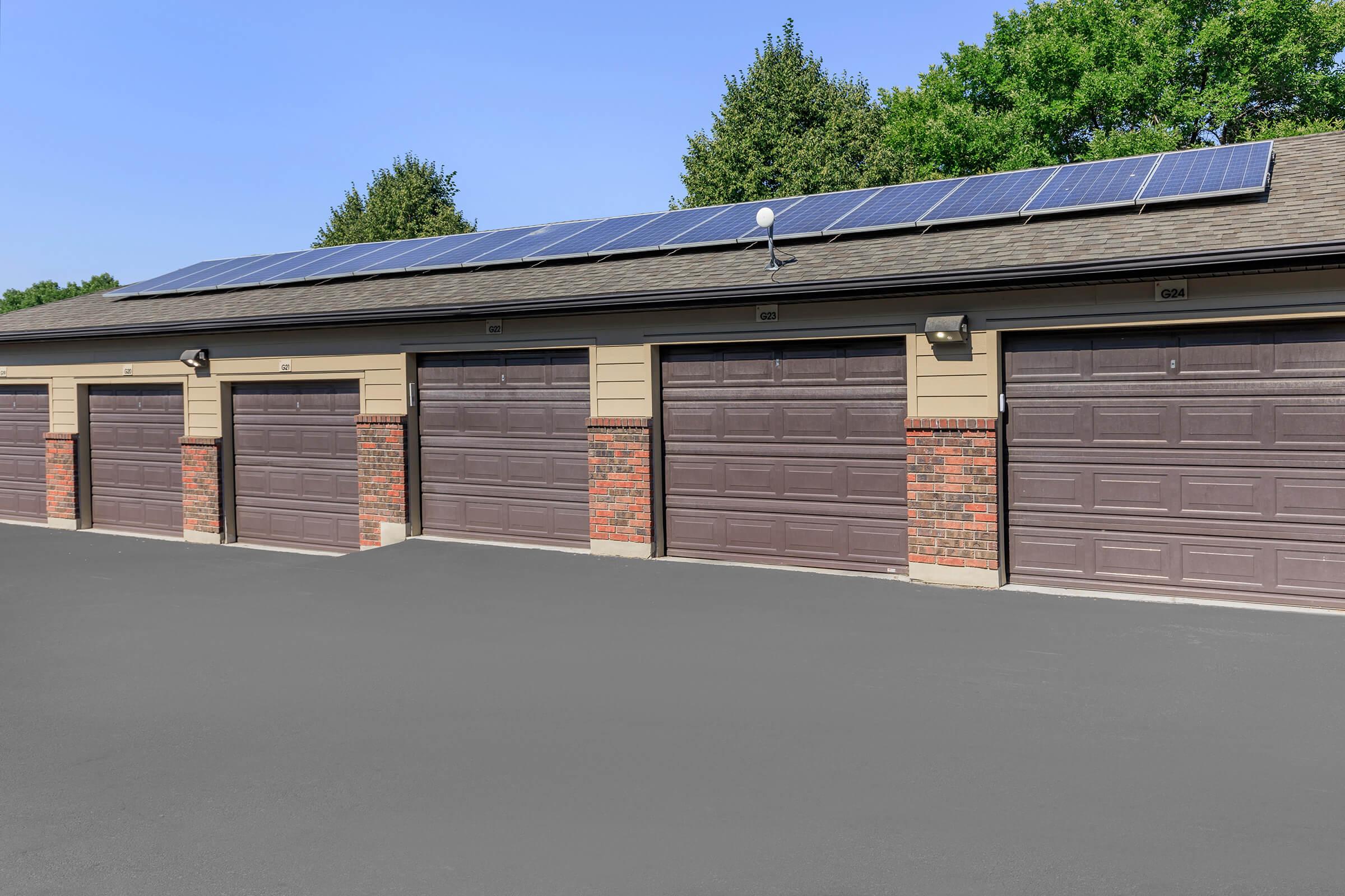 A row of garage doors, featuring dark brown doors with brick accents beneath them. Above, solar panels are installed on the roof. The background includes green trees and a clear blue sky, indicating a bright, sunny day.