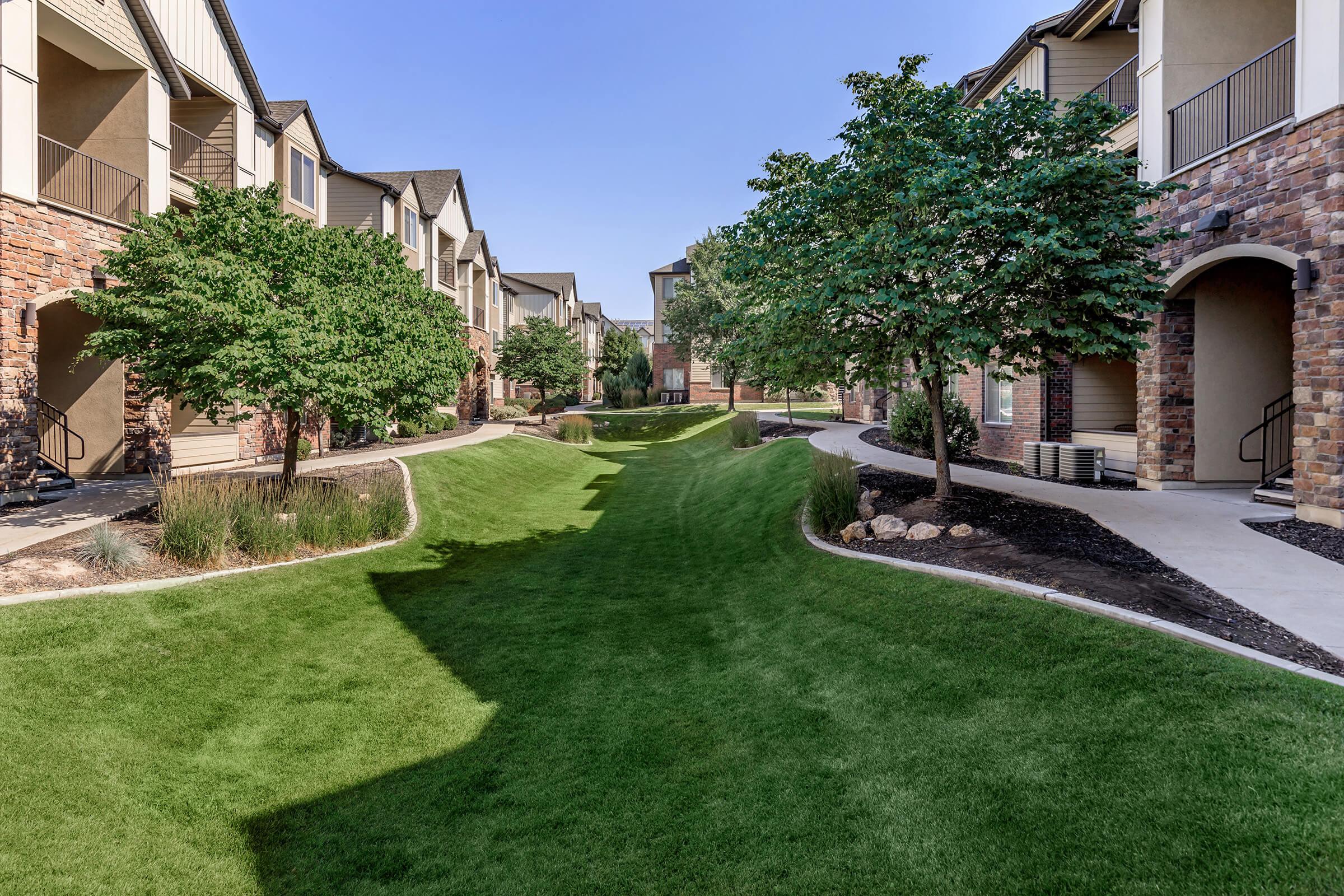 A spacious apartment community featuring landscaped green lawns and mature trees. Walkways meander through the area, leading to various building entrances. The scene is clear and sunny, highlighting the well-maintained grounds and the modern architecture of the buildings.