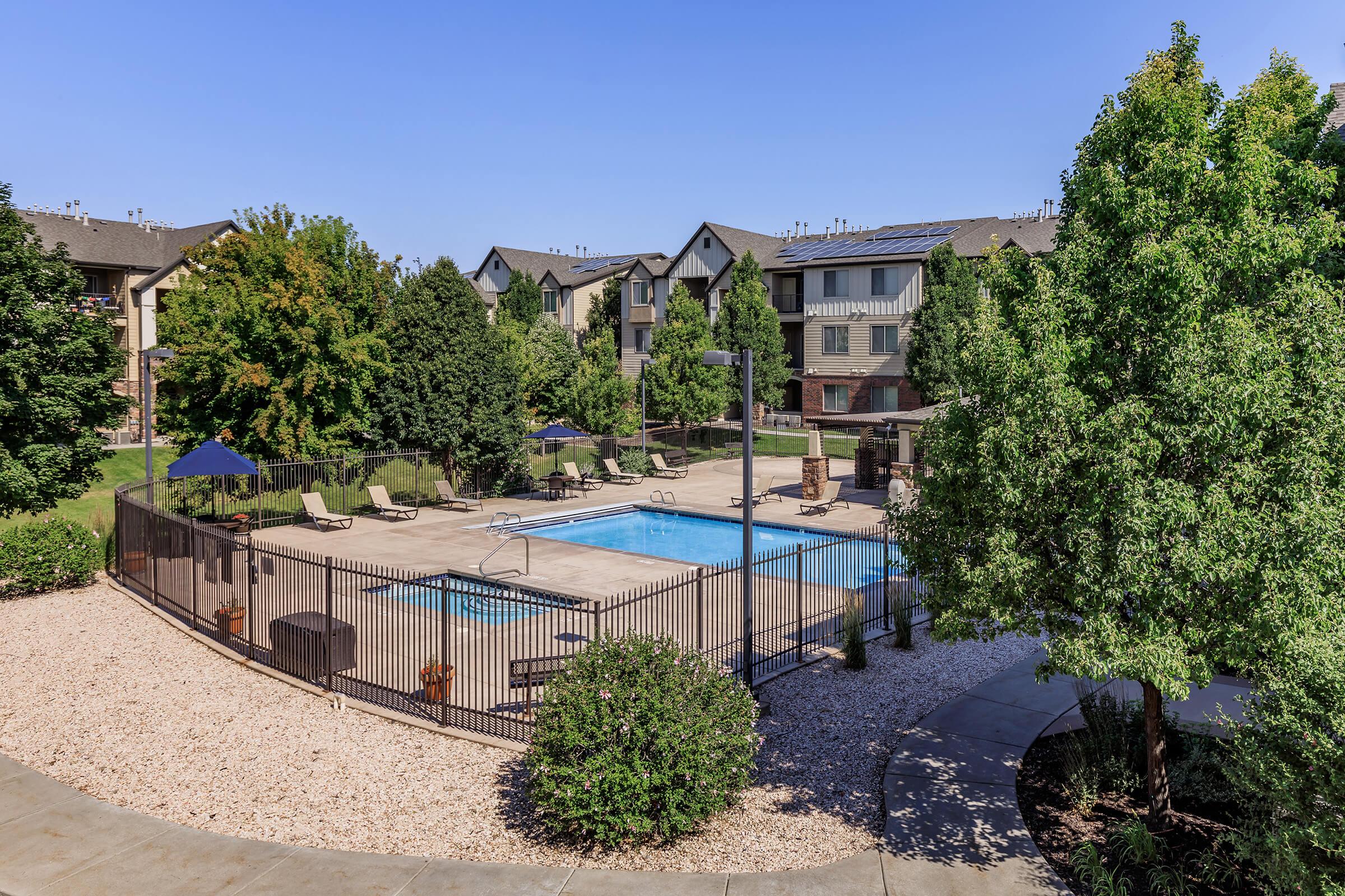 A serene outdoor pool area surrounded by lush greenery and residential buildings. The pool is enclosed by a black fence, with lounge chairs and umbrellas positioned nearby. The bright blue sky enhances the inviting atmosphere, perfect for relaxation and leisure.