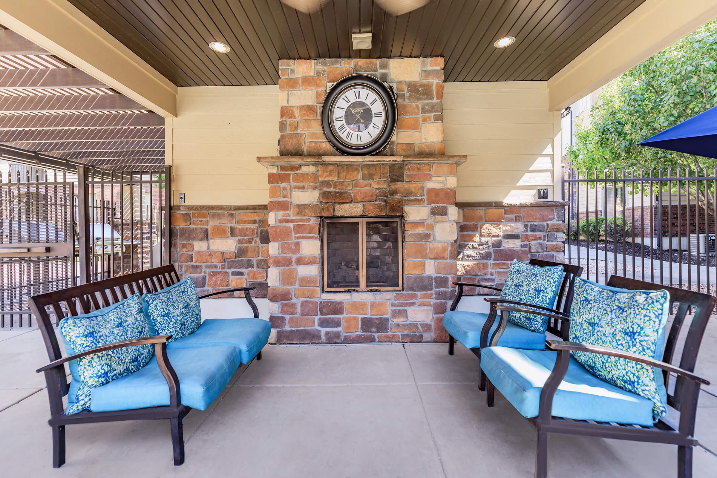 Outdoor lounge area featuring two wooden benches with blue cushions, positioned in front of a stone fireplace with a large clock mounted above it. The space is covered, with a ceiling that has wooden beams, creating a cozy atmosphere. Lush greenery is visible in the background.