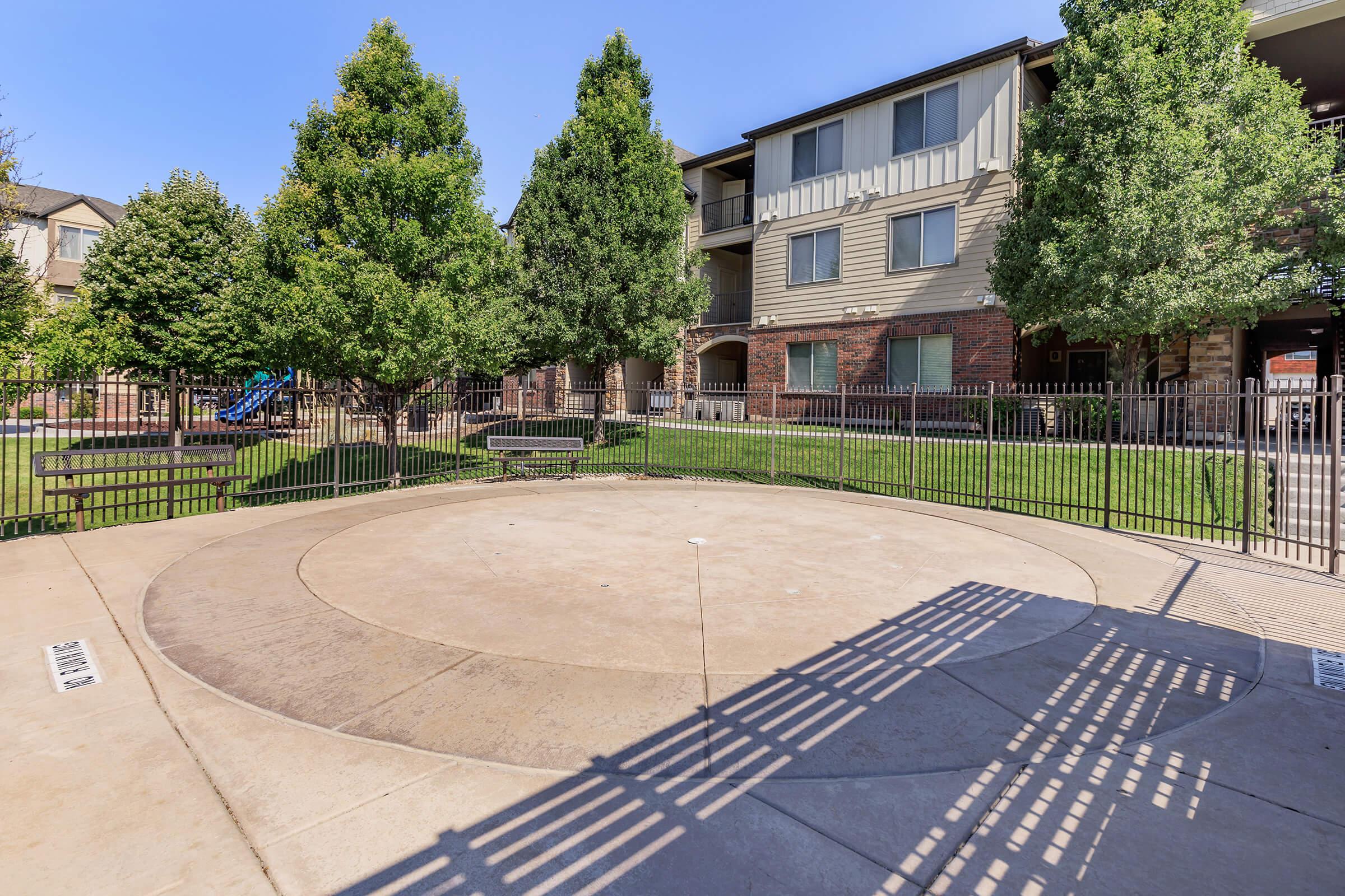 A circular concrete area surrounded by a fence, with green grass and trees in the background. There are benches along the perimeter and a multi-story apartment building in the background, showing a playground structure in the distance. The sky is clear and blue.