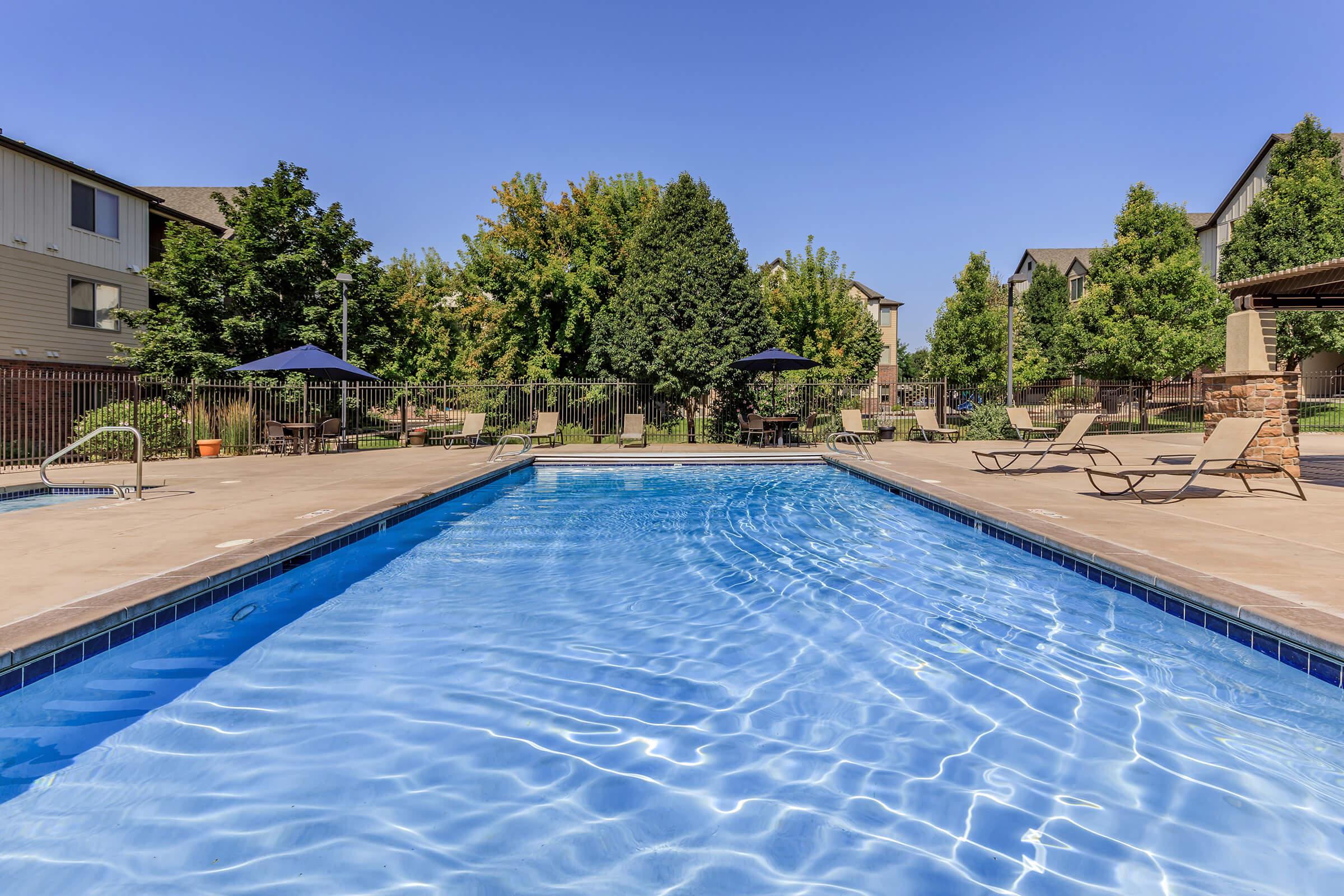 A clear blue swimming pool surrounded by loungers and umbrellas, set in a landscaped area with trees and residential buildings in the background. The sky is bright and blue, creating a peaceful and inviting atmosphere.