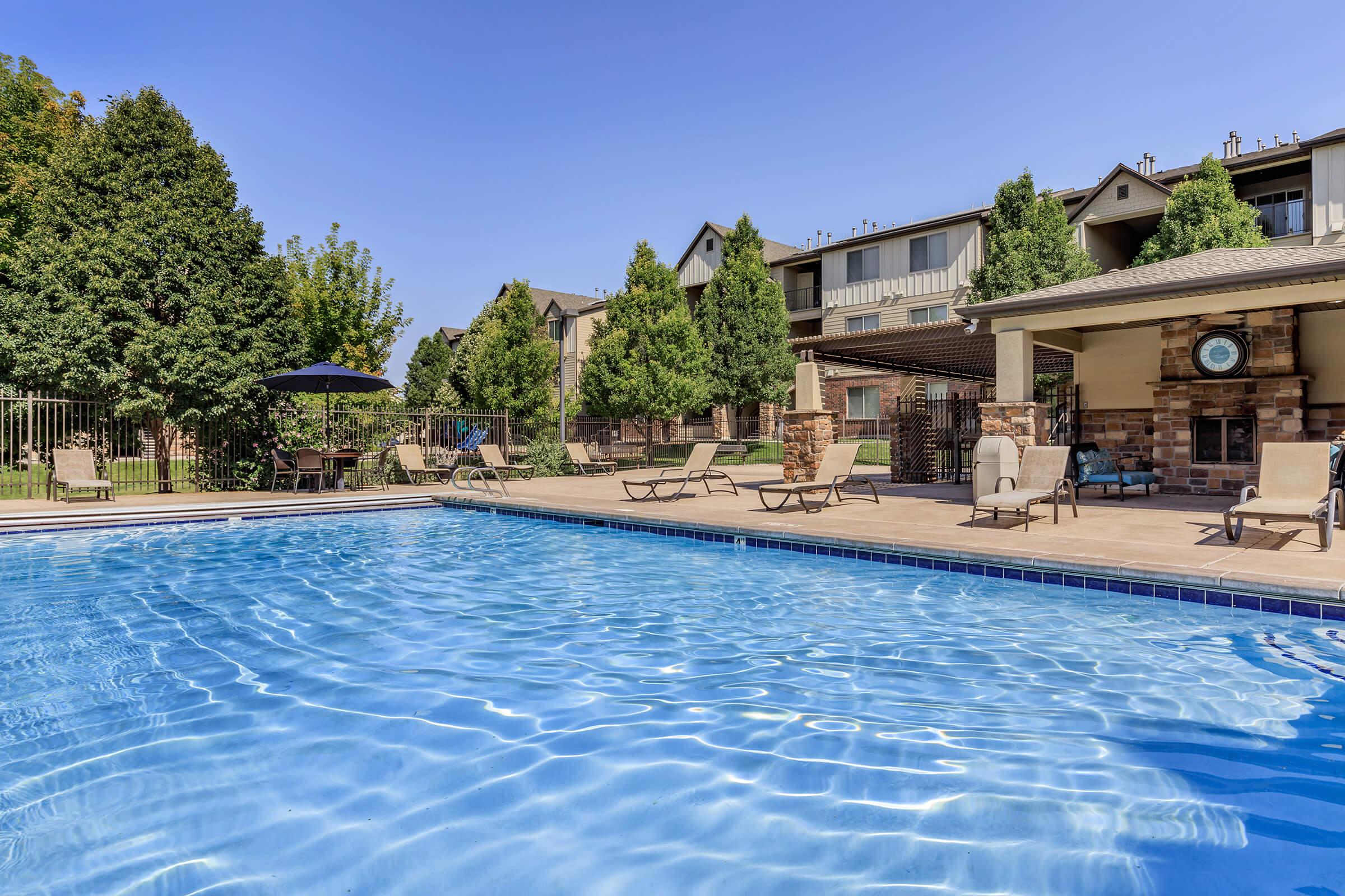 A clear blue swimming pool surrounded by lounge chairs and an umbrella. Lush green trees provide shade in the background, and there are residential buildings adjacent to the pool area. The sunny sky enhances the inviting atmosphere of the outdoor space.