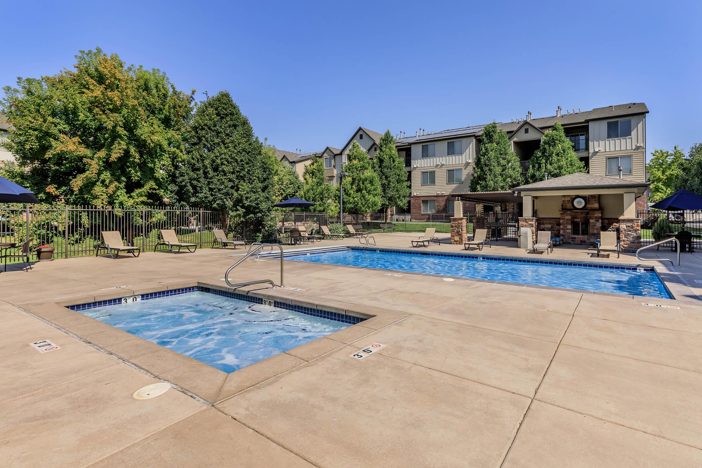 Outdoor pool area featuring a large swimming pool and a hot tub. Surrounding the pool are lounge chairs and shaded umbrellas, with green trees and apartment buildings in the background. The area is well-maintained and provides a relaxing environment for residents. Clear blue sky overhead.