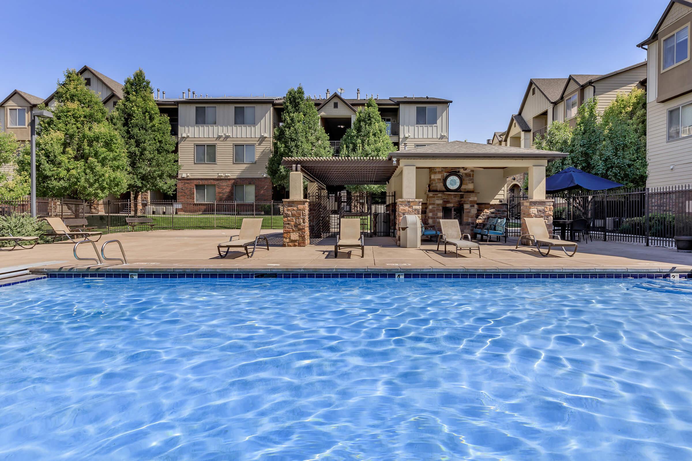 A swimming pool with clear blue water in the foreground, surrounded by lounge chairs and a stone patio. In the background, there is a multi-story apartment building with green trees and a pergola area providing shade. The scene is under a clear blue sky.