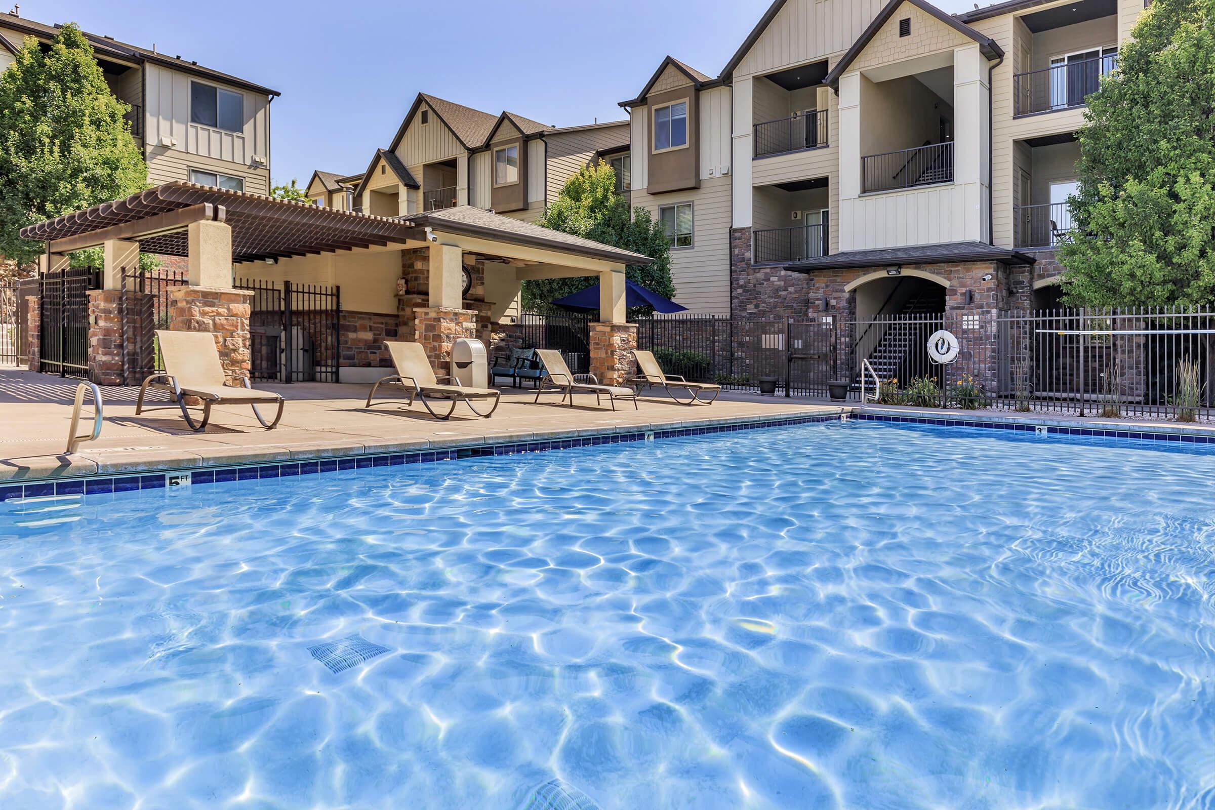 A serene outdoor swimming pool surrounded by lounge chairs, with a shaded seating area featuring a pergola. The backdrop consists of multi-story apartment buildings with balconies and lush greenery. The water sparkles under a clear blue sky, creating a tranquil ambiance.