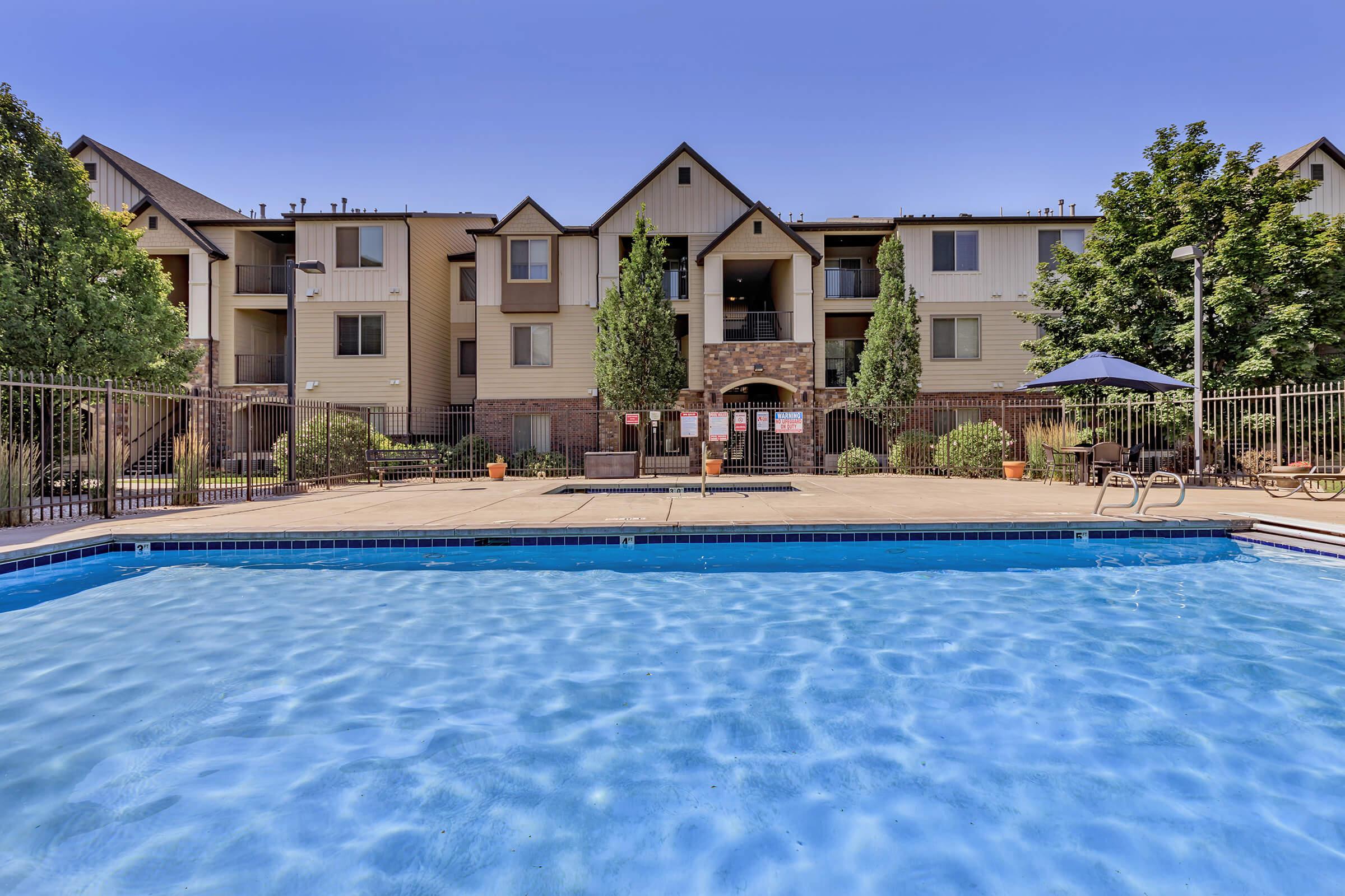 A clear swimming pool in the foreground with a landscaped area and an apartment building in the background. The building features multiple stories with a mix of windows and balconies, surrounded by green trees and shrubs. A lounge area with chairs is visible near the pool.