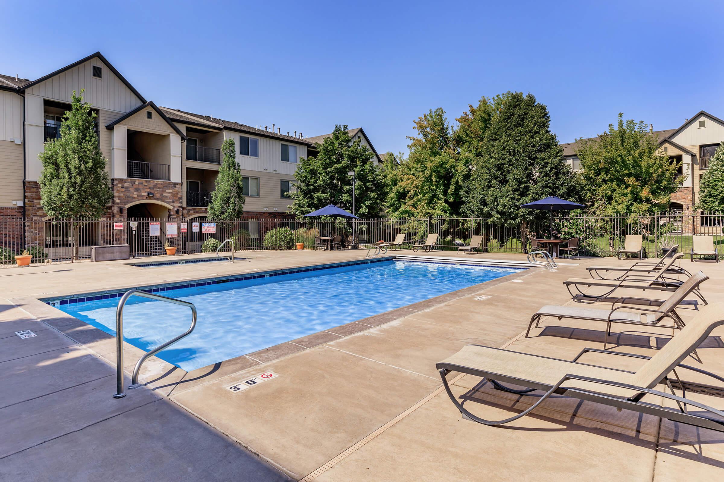 A clear blue swimming pool surrounded by lounge chairs, with umbrellas for shade. In the background, there are multi-story apartment buildings and lush greenery, creating a serene outdoor space perfect for relaxation. It’s a sunny day with a bright blue sky.