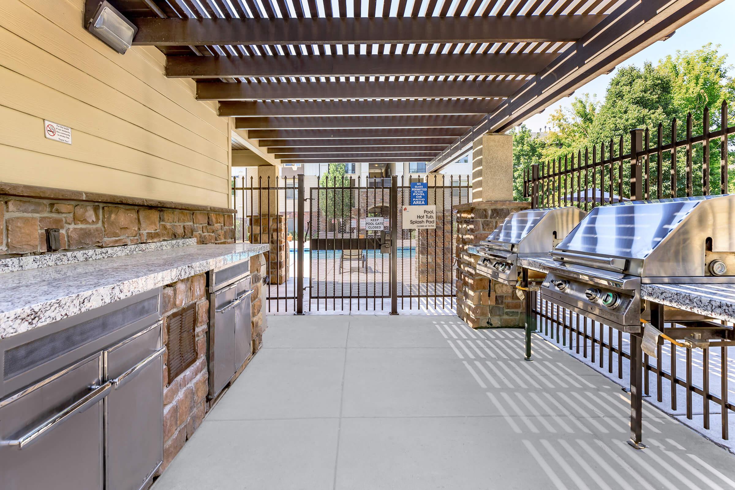 Outdoor grilling area with two stainless steel grills and a granite countertop. The space is shaded by a pergola, with a fenced pool area visible in the background. Sunlight creates shadows on the ground, enhancing the inviting atmosphere for gatherings and barbecues.