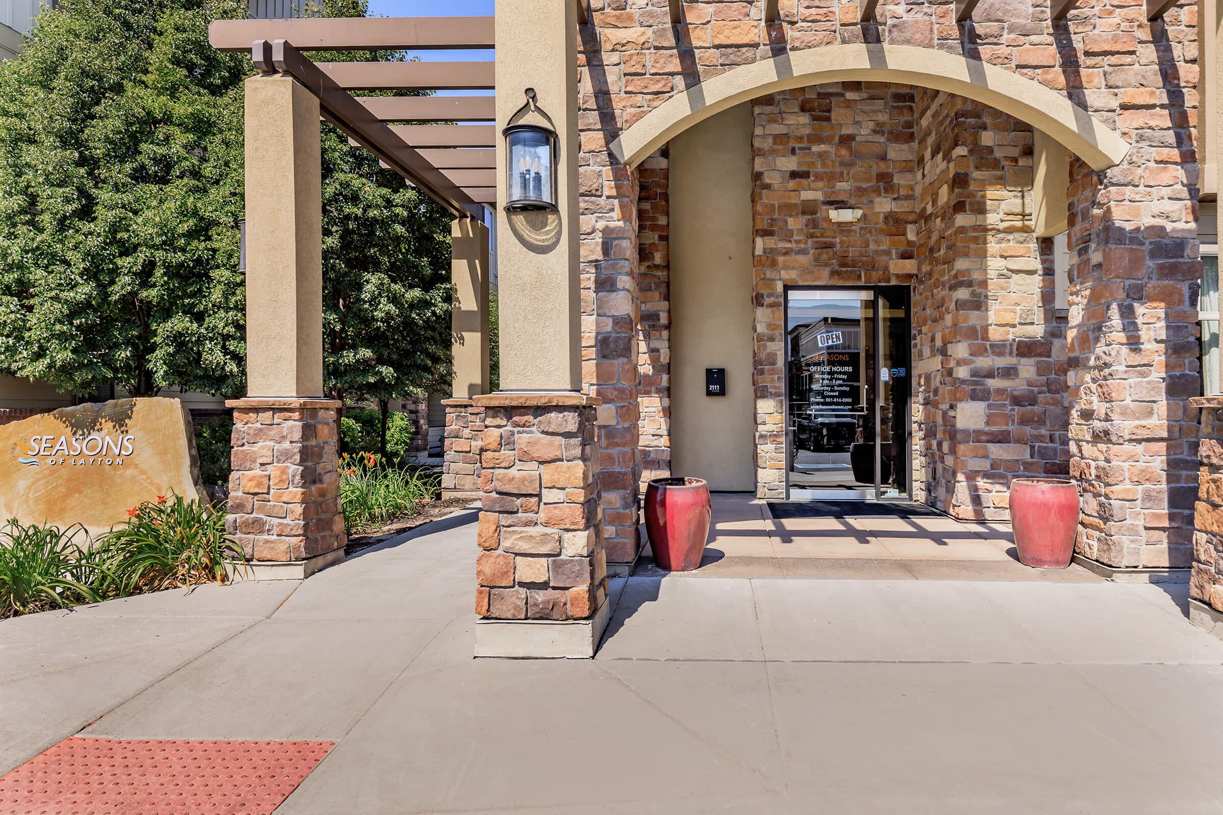 Entrance of a building featuring a stone façade, large archway, and decorative lighting. To the left, a sign reading "Seasons" is displayed, with vibrant red planters on either side of the entrance. The pathway is paved, leading up to double glass doors that invite visitors inside. Lush greenery is visible in the background.