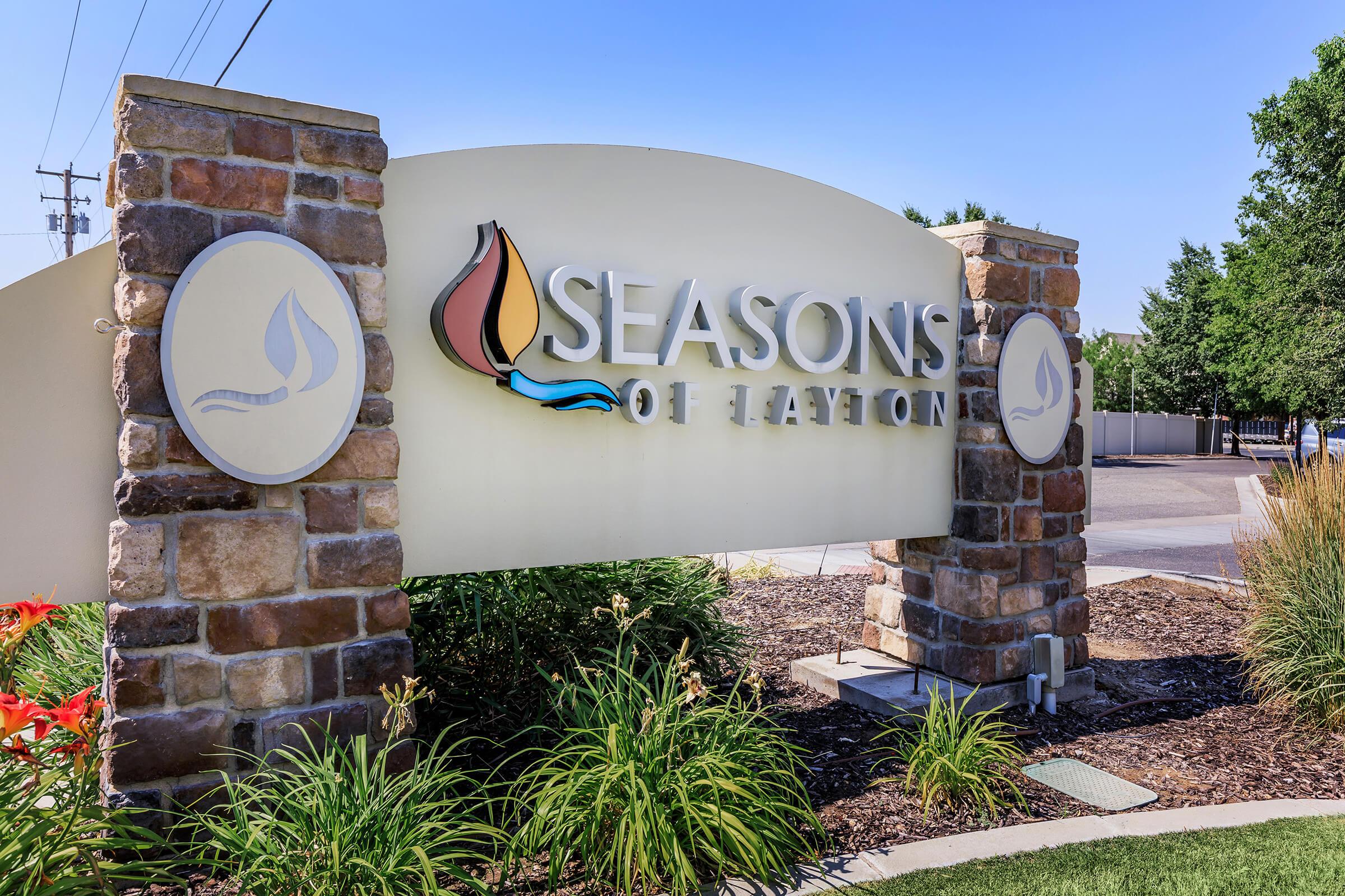 A decorative sign for "Seasons of Layton," featuring a stylized water droplet and flame logo. The sign is made of stone and has vibrant landscaping in the foreground, set against a clear blue sky.