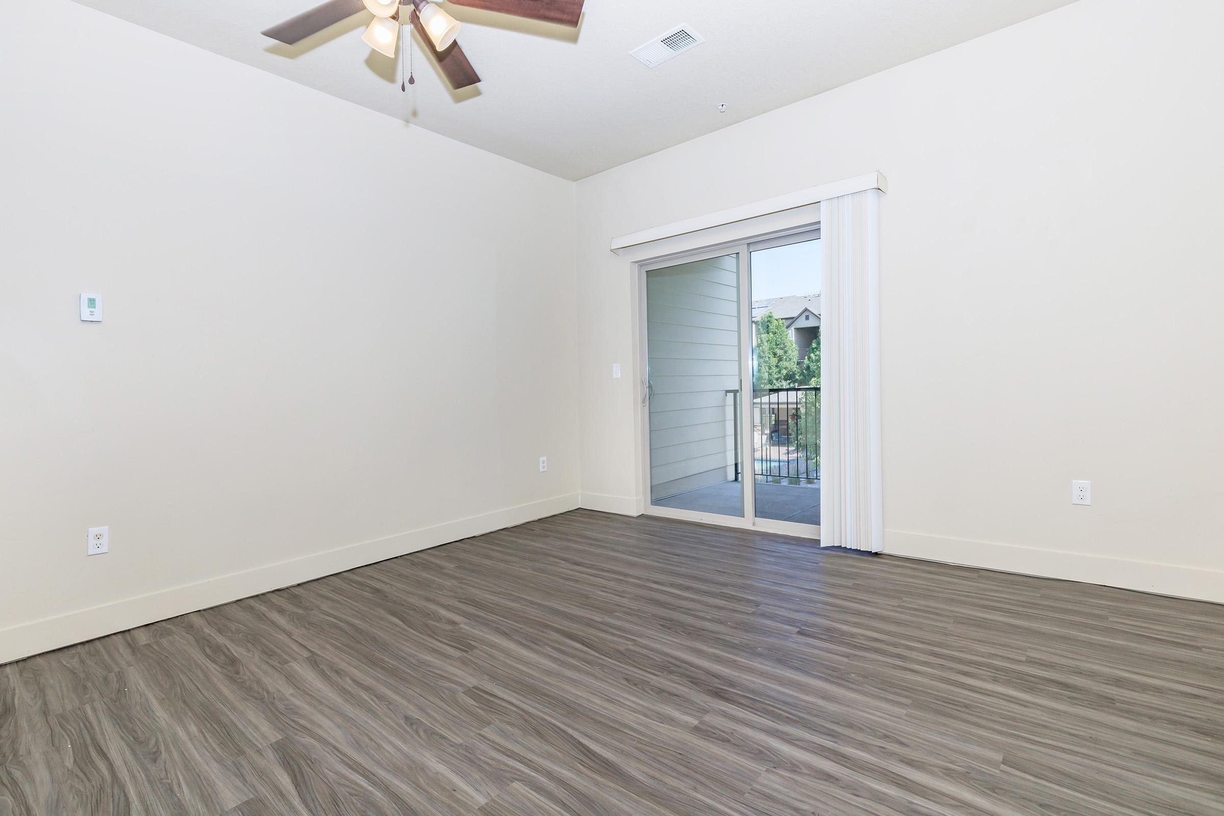 A spacious, empty room with light-colored walls and a ceiling fan. The floor features modern laminate planks. Large sliding glass doors lead to a balcony with outdoor views, allowing natural light to fill the space.