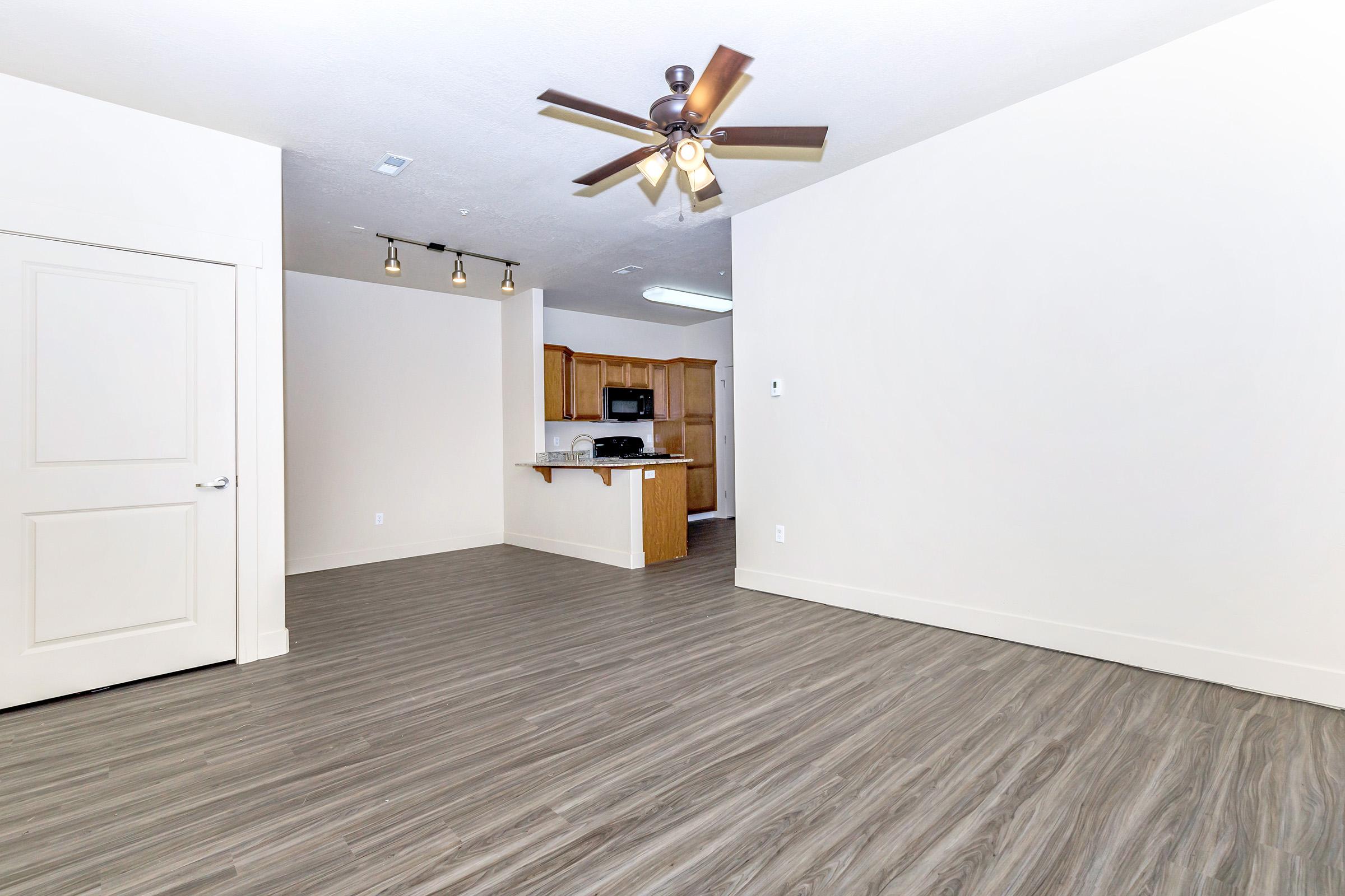 A spacious, modern living room featuring light wood-look flooring, a ceiling fan, and a doorway leading to a kitchen with wooden cabinets and appliances visible in the background. The walls are painted a neutral color, creating a clean and inviting atmosphere.