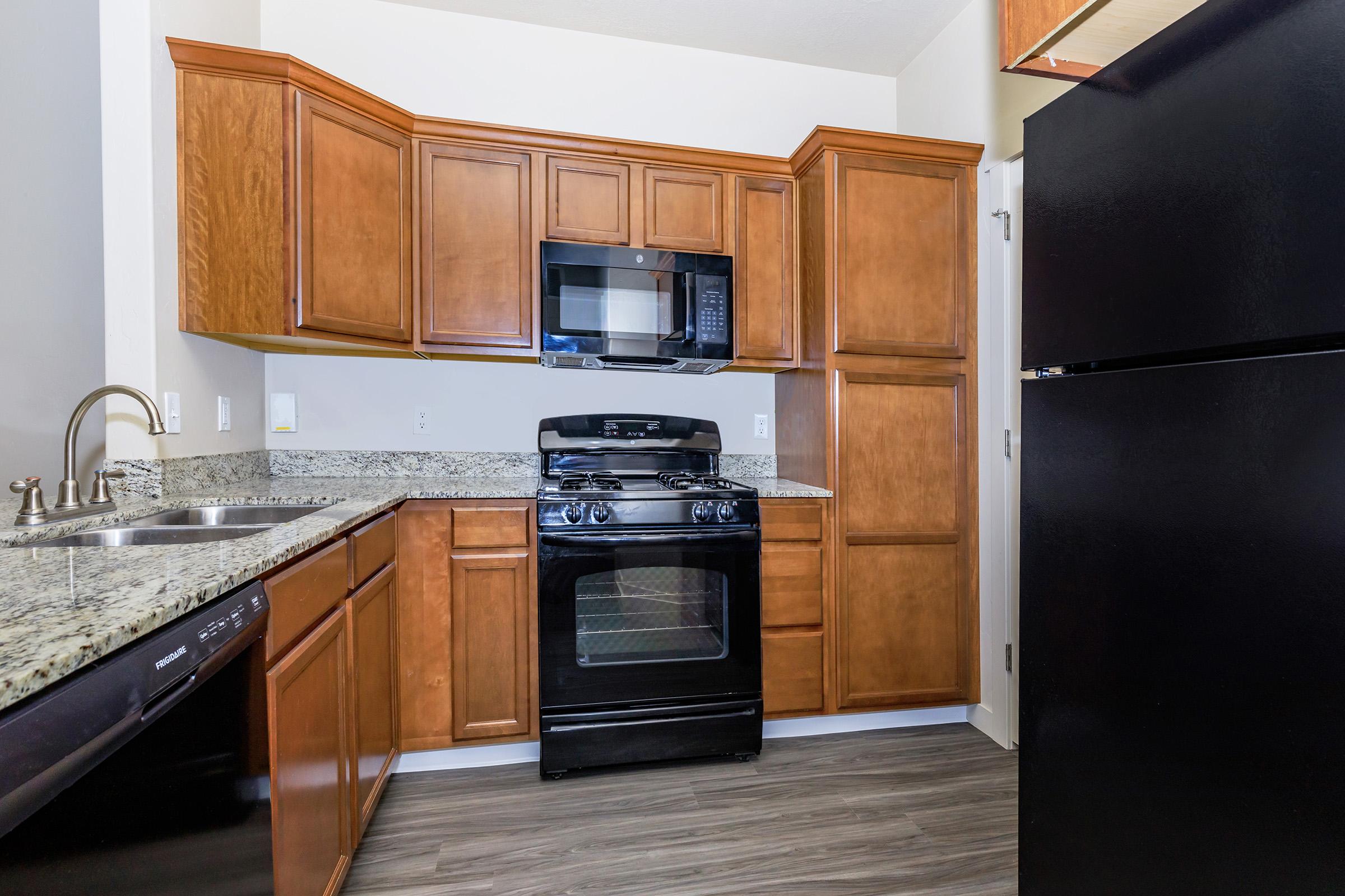 A modern kitchen featuring wooden cabinets, a granite countertop, a stainless steel sink, a black gas stove, a microwave, and a black refrigerator. The kitchen has a sleek, contemporary design with warm wood tones and a clean layout.