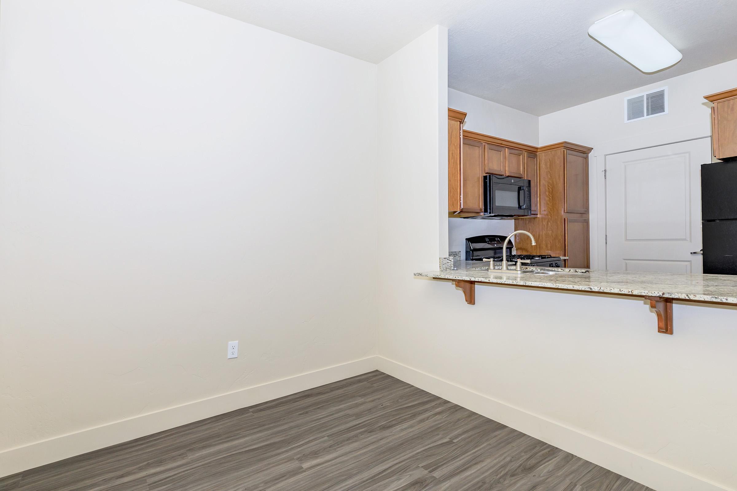 A modern kitchen with wooden cabinets and a granite countertop. The space features a black microwave and an oven, with a view into an open area. The flooring is a light wood finish, and the walls are painted in a neutral color. There's a small bar or ledge with a sink integrated into the countertop.