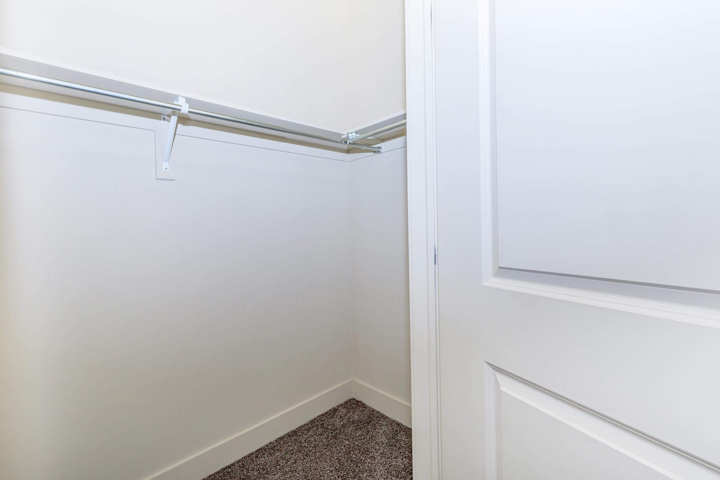 A partially empty closet with white walls and a single shelf. The closet features a door slightly ajar, revealing a carpeted floor. The shelves are mounted on the left side, creating an open and spacious area ideal for storage. The overall appearance is clean and minimalistic.