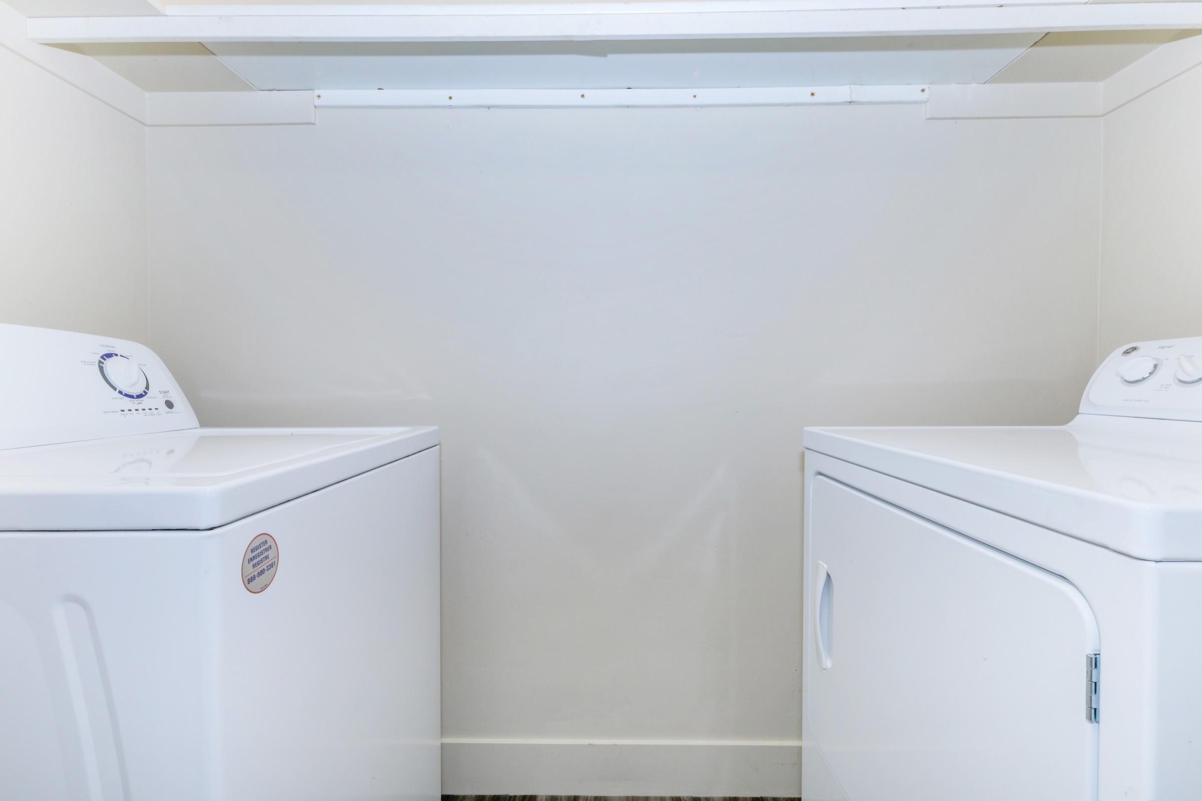 A clean laundry room featuring a white washing machine on the left and a white dryer on the right. The machines are positioned against a plain, light-colored wall with a shelf above, creating a simple and organized appearance. The floor is a light-colored material.