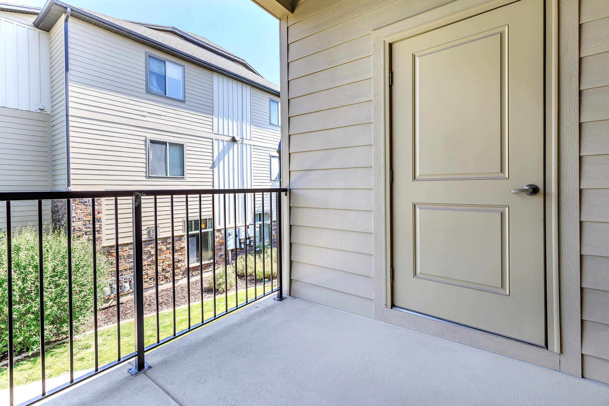 View of a balcony with a closed beige door leading to an apartment. The railing is visible, and in the background, there are neighboring apartment buildings with exterior siding and windows. The ground is covered with grass and landscaping, suggesting a residential area.