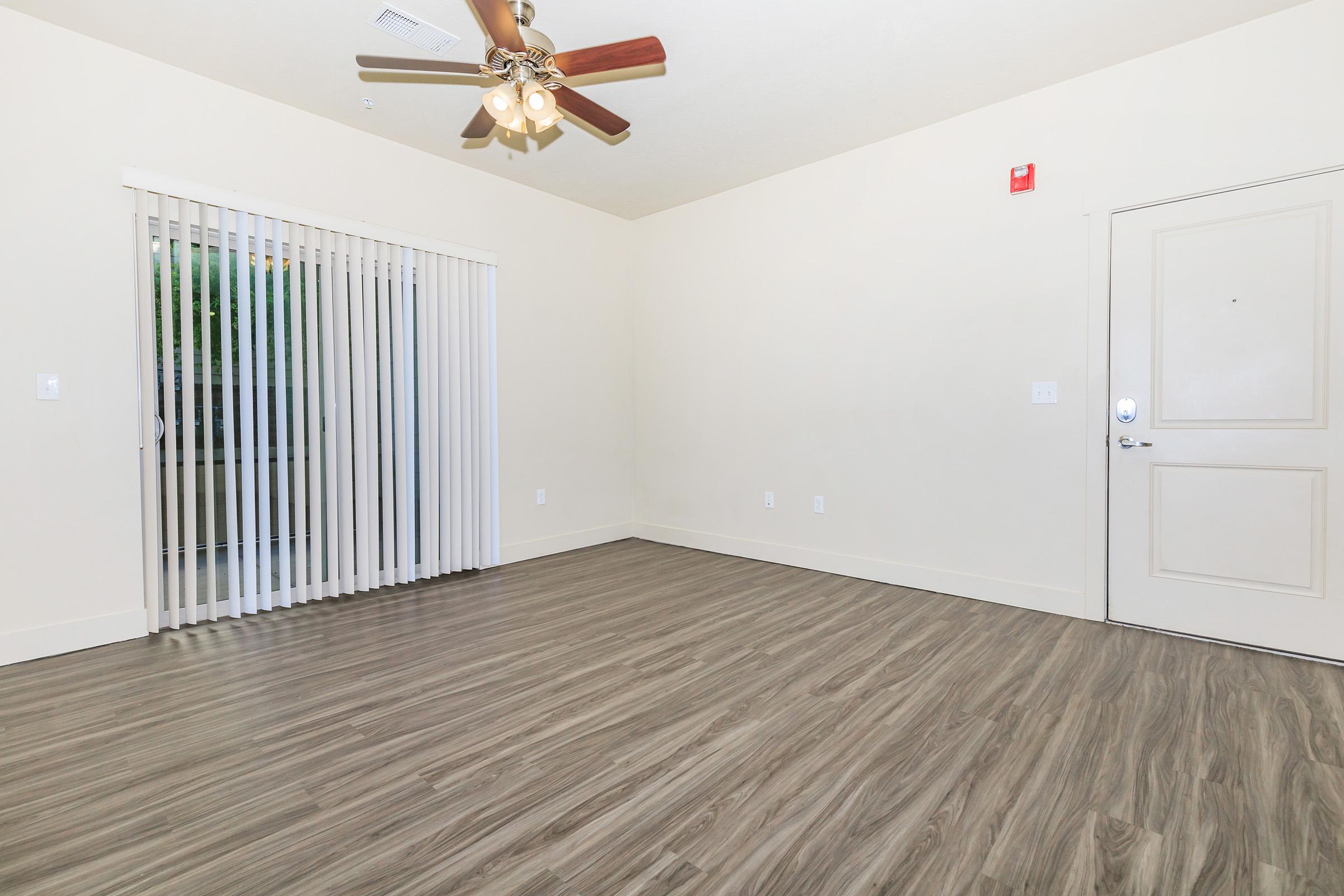 Empty room with light-colored walls and a ceiling fan. It features a sliding glass door with vertical blinds leading to an outdoor space. The floor has a wood-like finish, and there’s a light switch and a door on one wall. The overall appearance is spacious and airy.