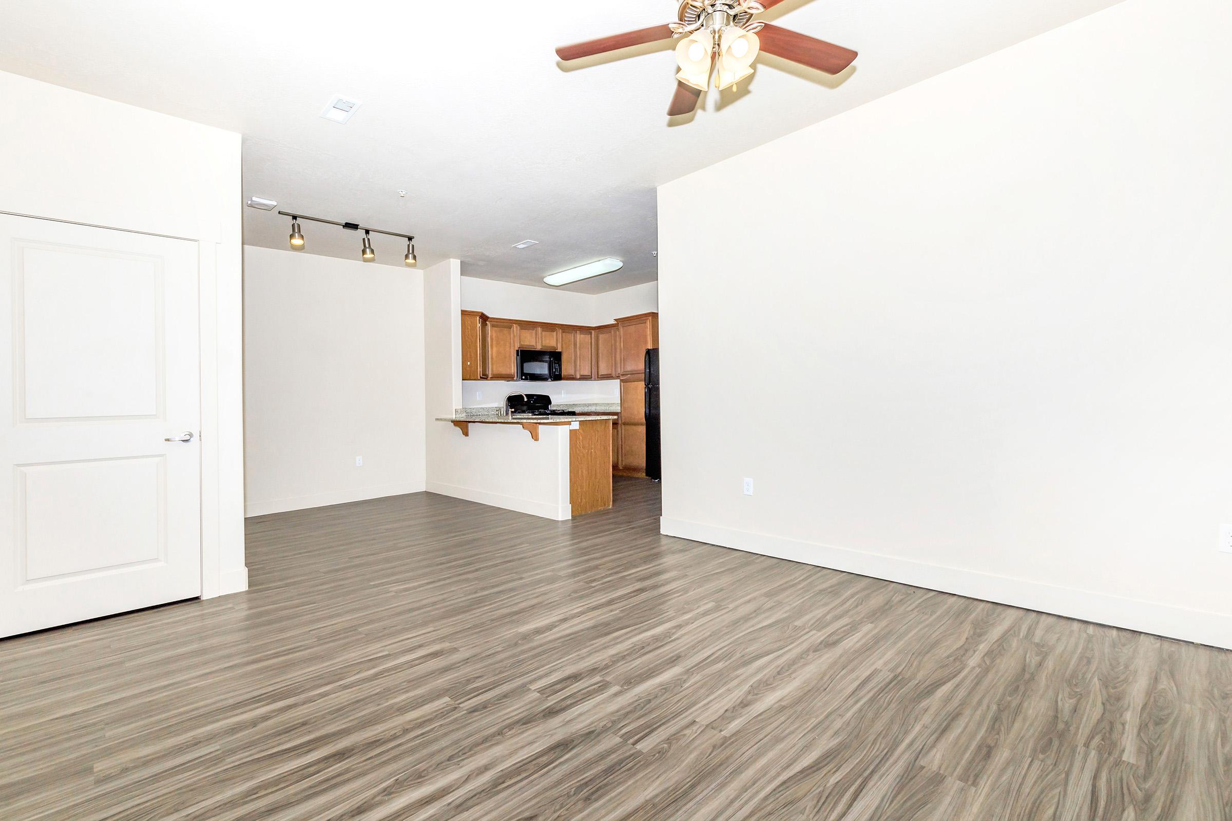 A spacious living area with light-colored walls and modern wood-like flooring. There is a ceiling fan, and in the background, a kitchen with wooden cabinets and stainless steel appliances is visible. The open layout creates a bright and inviting atmosphere.