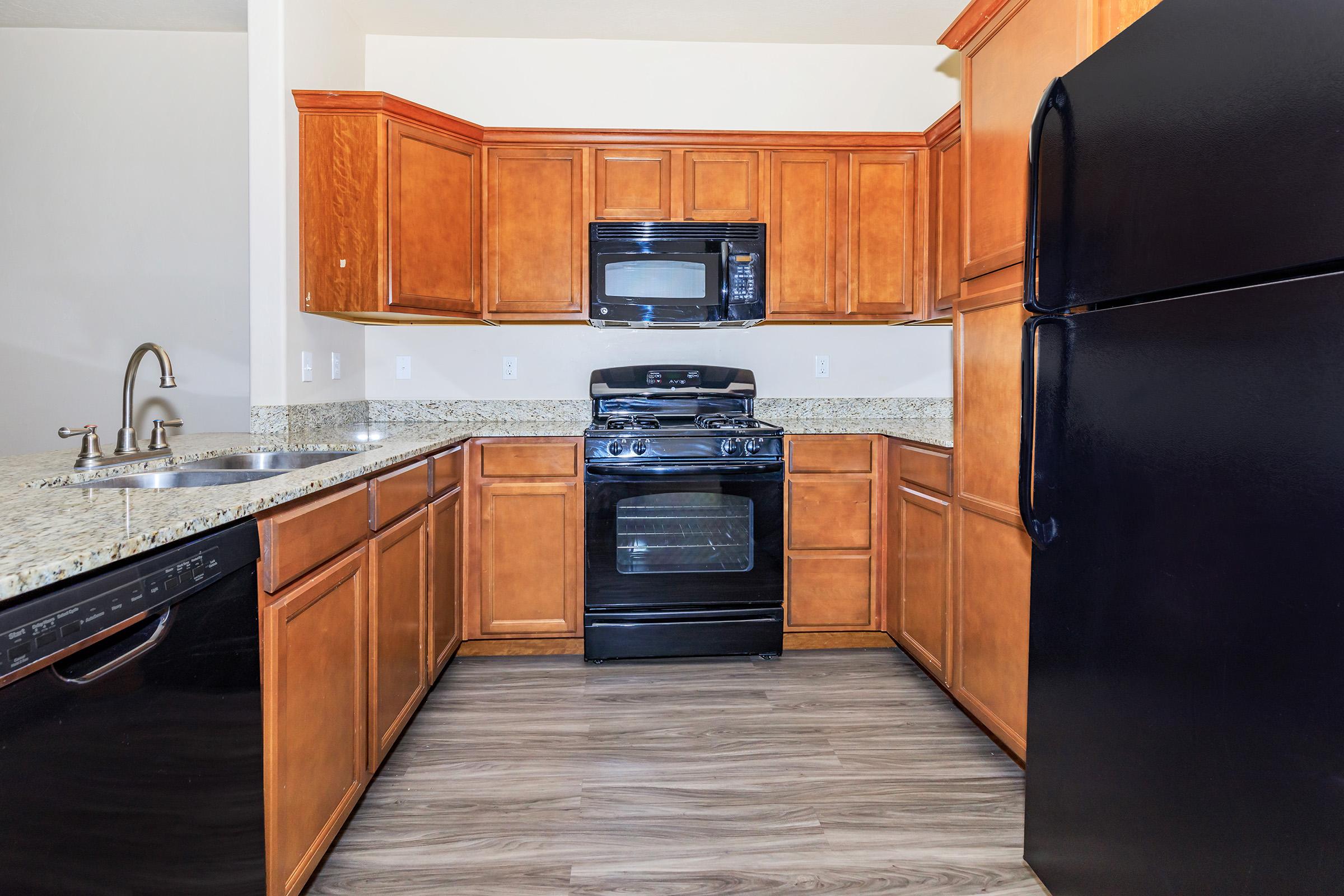 Modern kitchen featuring wooden cabinets, a black refrigerator, a black gas stove, and a built-in microwave. The countertops are made of granite, and there is a double sink on the left. The flooring is a light-colored laminate, creating a warm and inviting atmosphere.