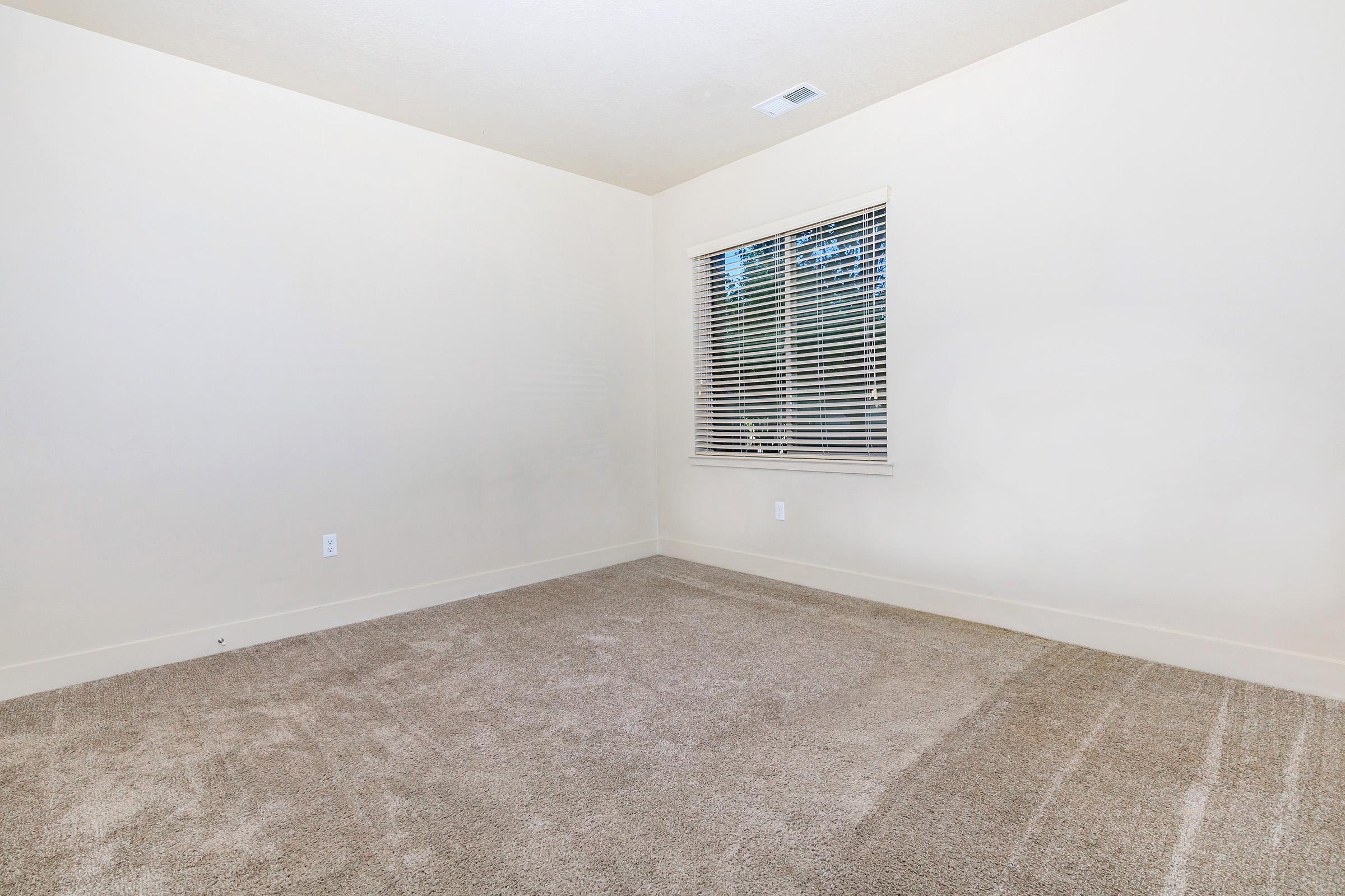 A spacious, empty room with light beige walls and a large window covered by white blinds. The floor is carpeted in a light brown color, and there are no furniture or decorations in the space, creating a minimalist and airy atmosphere.