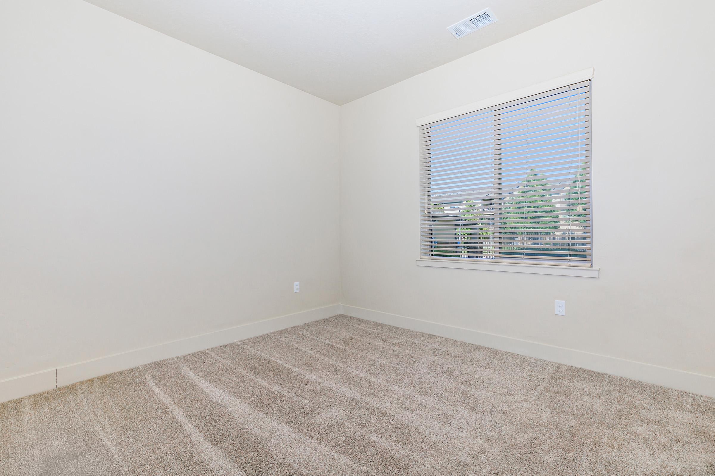 A small, empty room with beige walls and light-colored carpet. A window with blinds allows natural light to enter, showcasing a view of trees outside. The room has no furniture, creating a spacious and minimalist appearance.