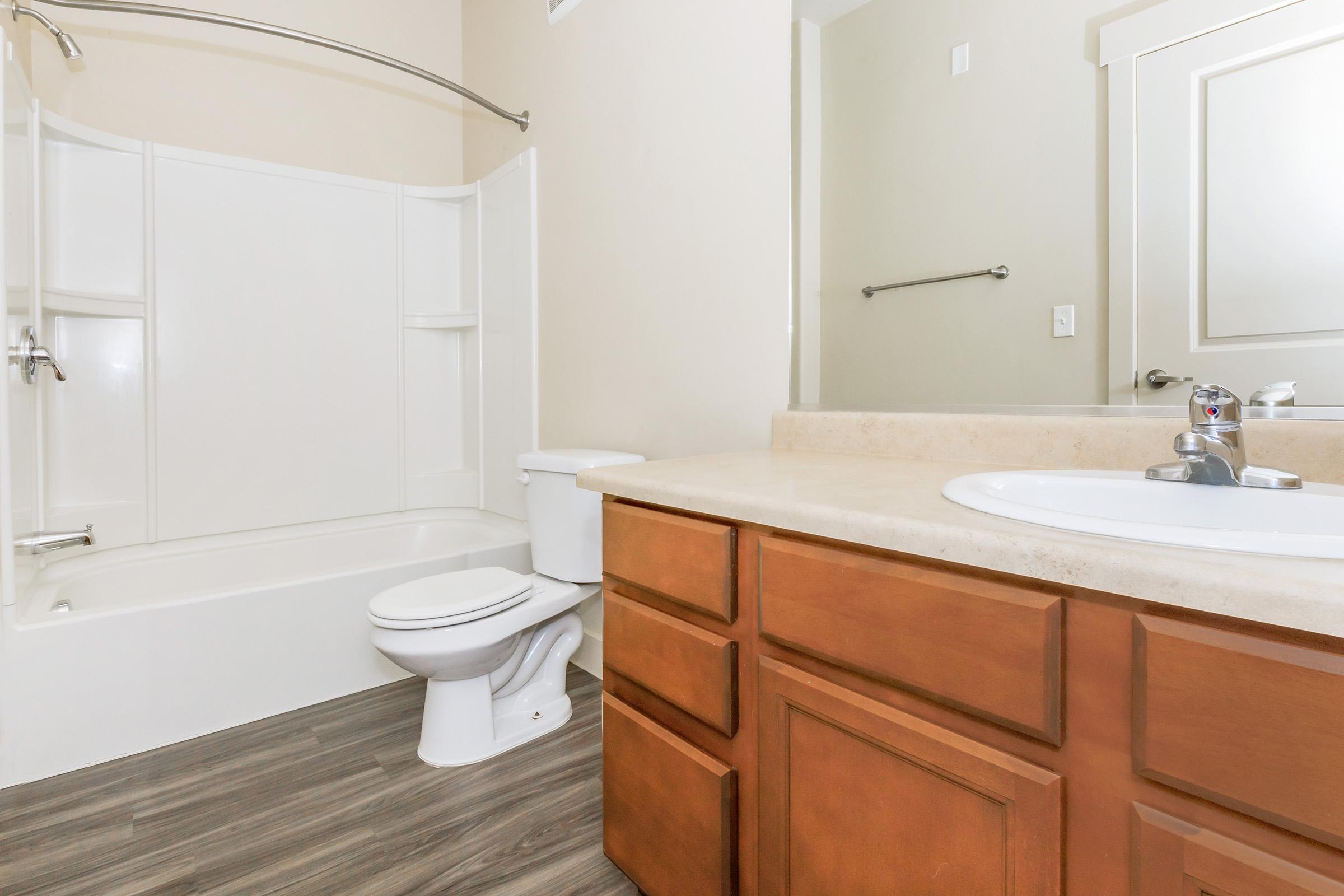 A modern bathroom featuring a white bathtub with a shower, a toilet, and a wooden vanity with a sink. The walls are painted light beige, and there is a framed mirror above the sink. The flooring is gray, resembling wood. Bright lighting enhances the clean and spacious design of the room.