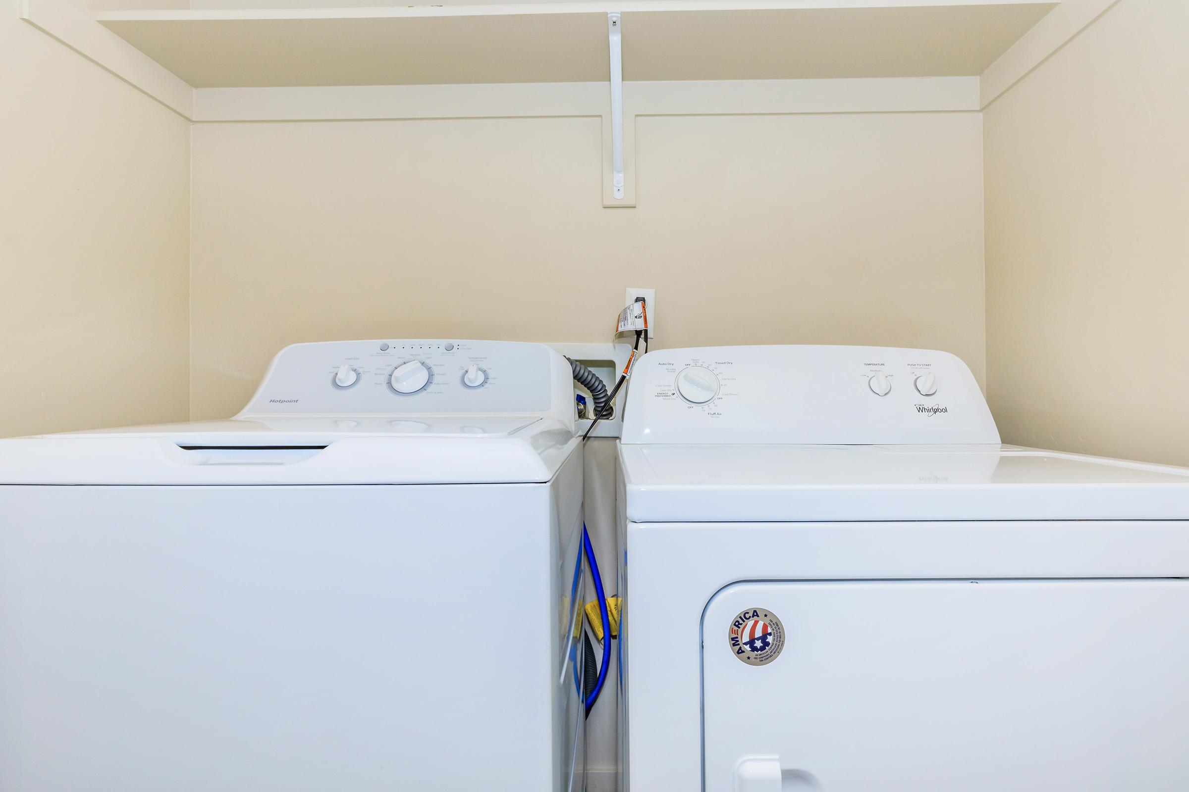 A white washing machine and a white dryer side by side in a laundry room, with a shelf above. The machines are against a light-colored wall, with a power outlet visible behind the washer. The washing machine has dials and knobs on the front.