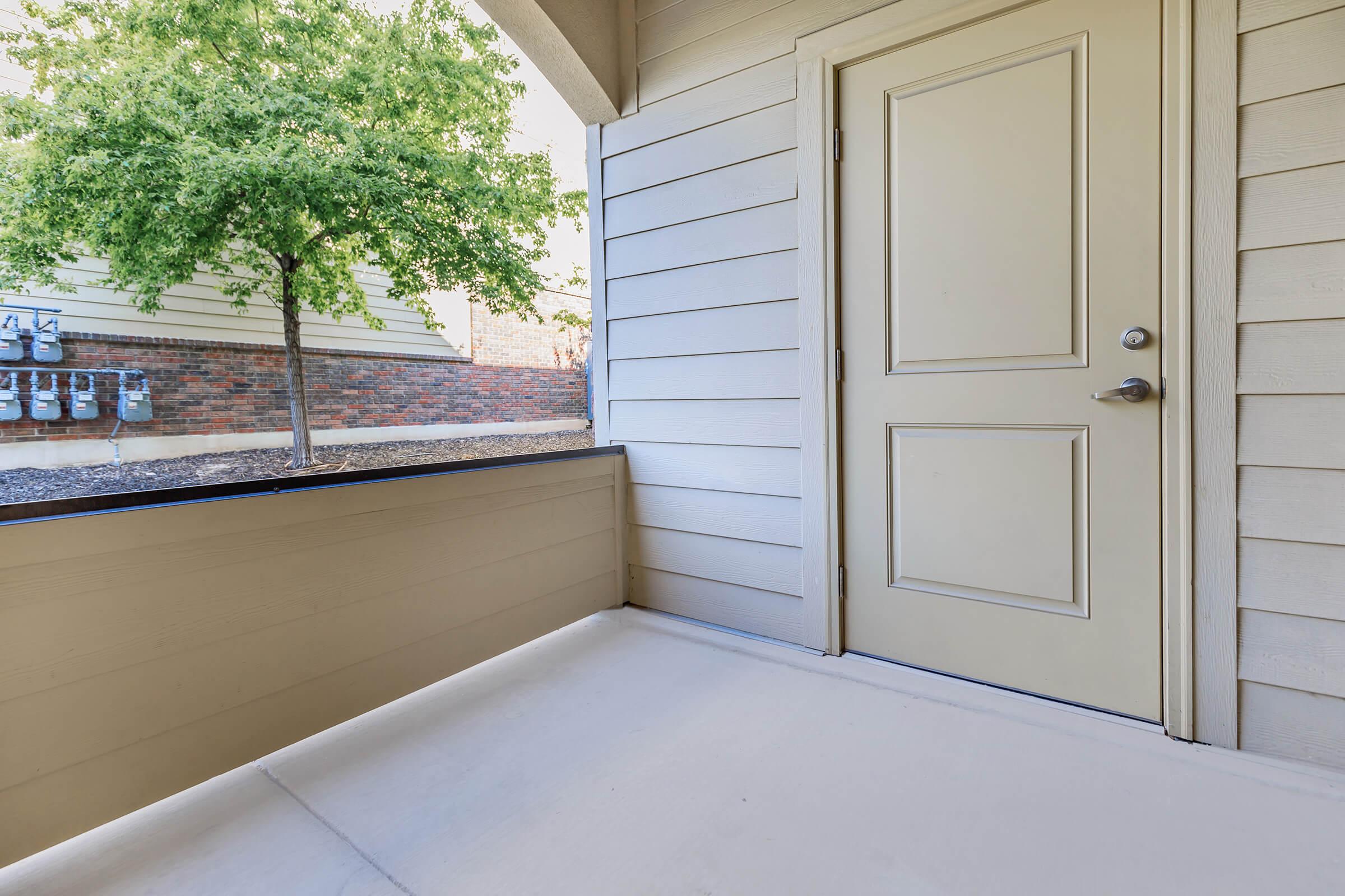 A well-lit entrance area featuring a beige door, with a small tree visible outside and several utility meters mounted on the wall nearby. The space has light-colored siding and a concrete floor, creating a clean and inviting entrance.